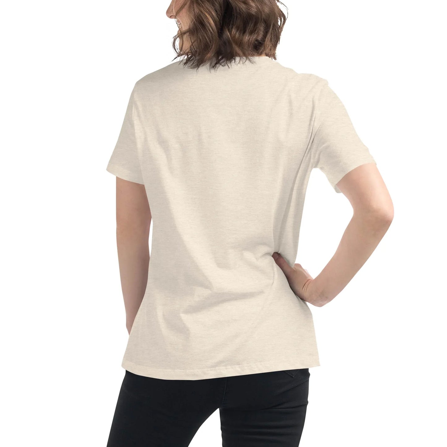 A person with wavy brown hair stands facing away, wearing a plain light beige Mambí t-shirt and black pants. One hand is on their hip, the other arm relaxed at their side. The background is plain white.