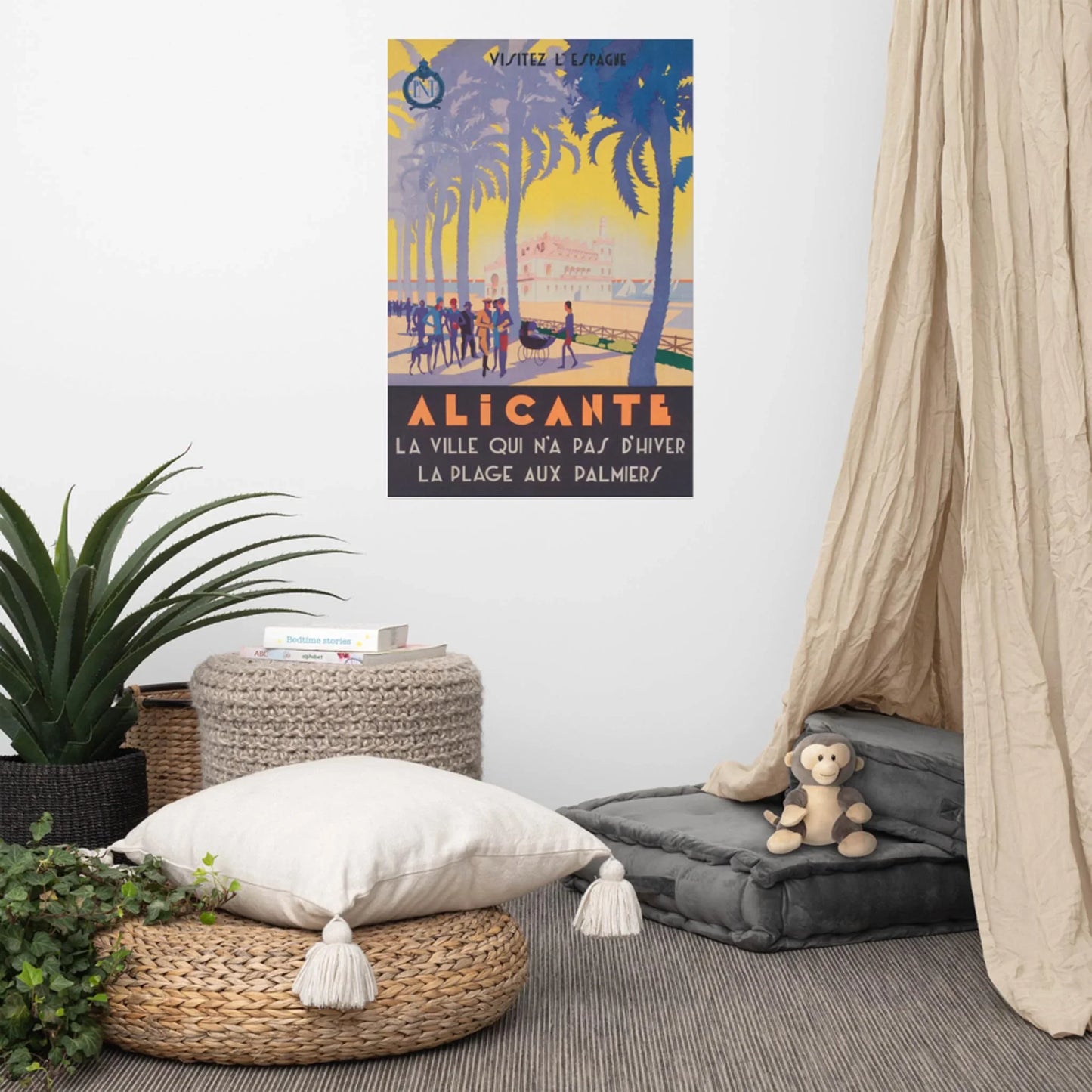 A cozy room corner with floor cushions, a plush toy, plants, woven baskets, and a Mambí touch. On the wall is a vintage-style poster of Alicante with palm trees and people, reading Alicante, la ville qui n’a pas d’hiver, la plage aux palmiers.