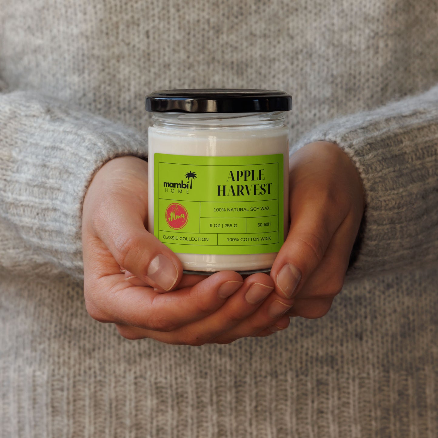 A person in a cozy gray sweater holds a glass jar candle labeled APPLE HARVEST—one of Mambi Homes eco-friendly candles, made with 100% natural soy wax and perfect for lovers of scented soy candles.