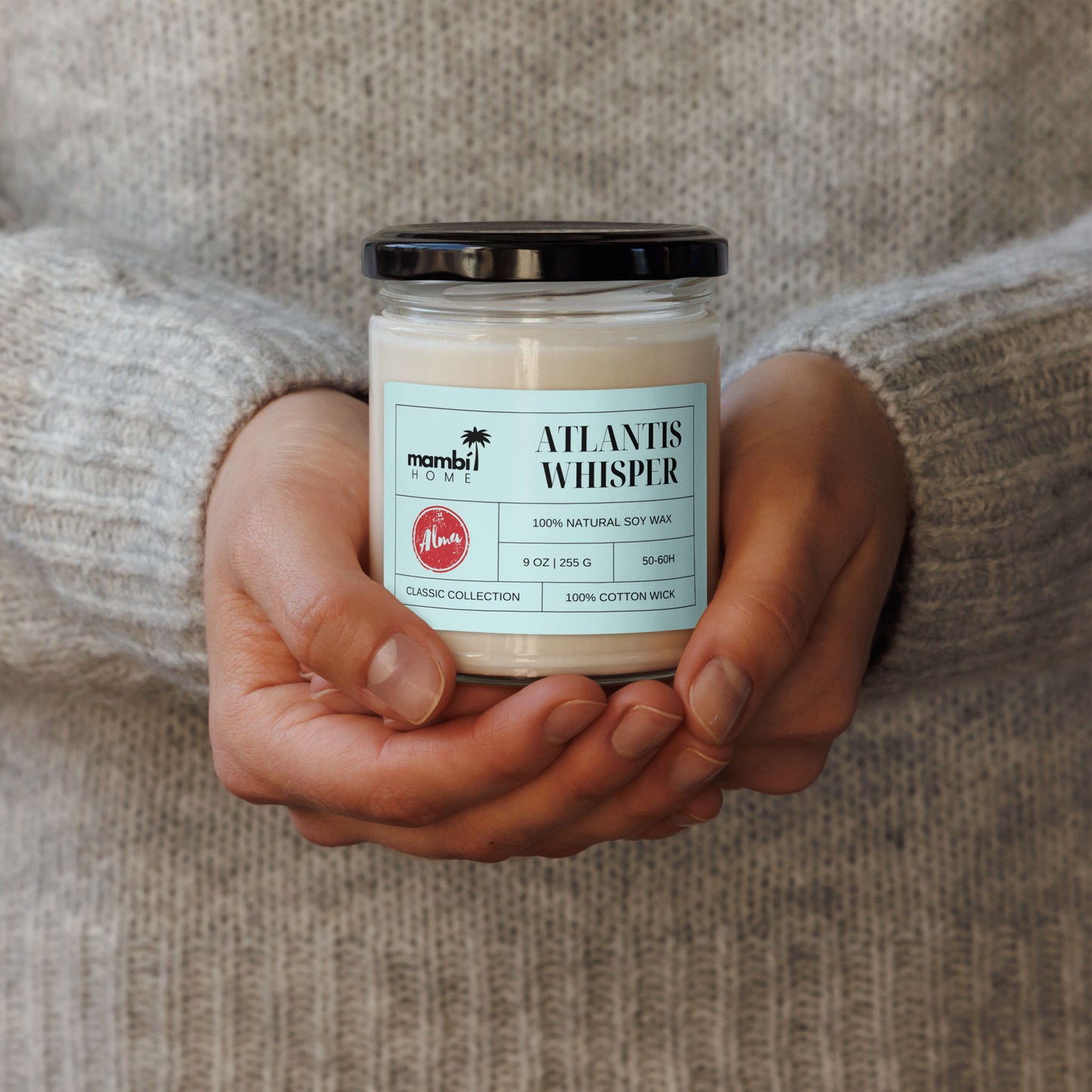 A person wearing a light grey sweater holds a glass jar candle labeled ATLANTIS WHISPER, made from 100% natural soy wax and 100% cotton wick—perfect for those who love natural soy wax candles.