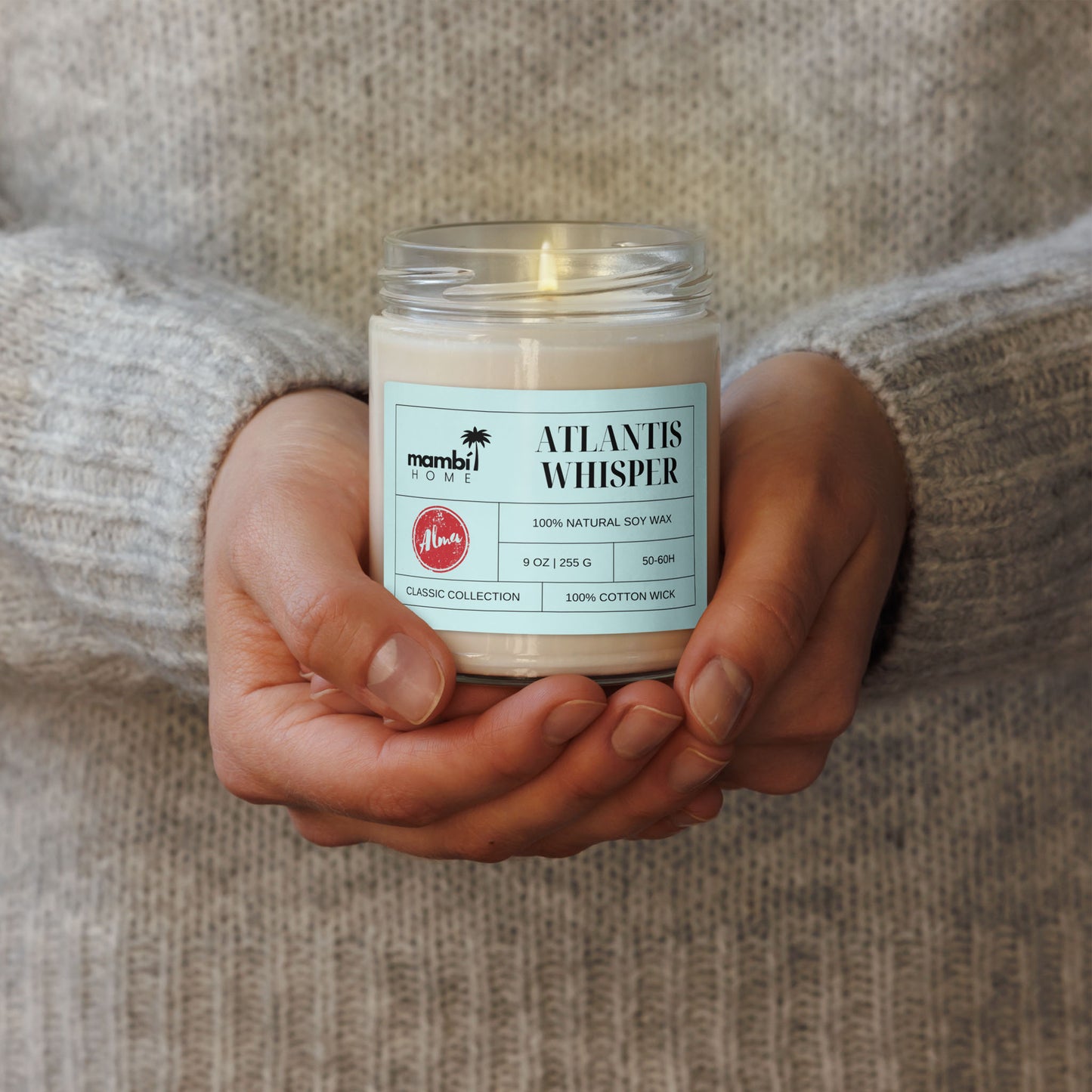 A person wearing a light gray sweater holds a jar labeled Atlantis Whisper with both hands. The label highlights palm tree graphics and describes it as one of their natural soy wax candles, 100% soy, 9 oz/255g.
