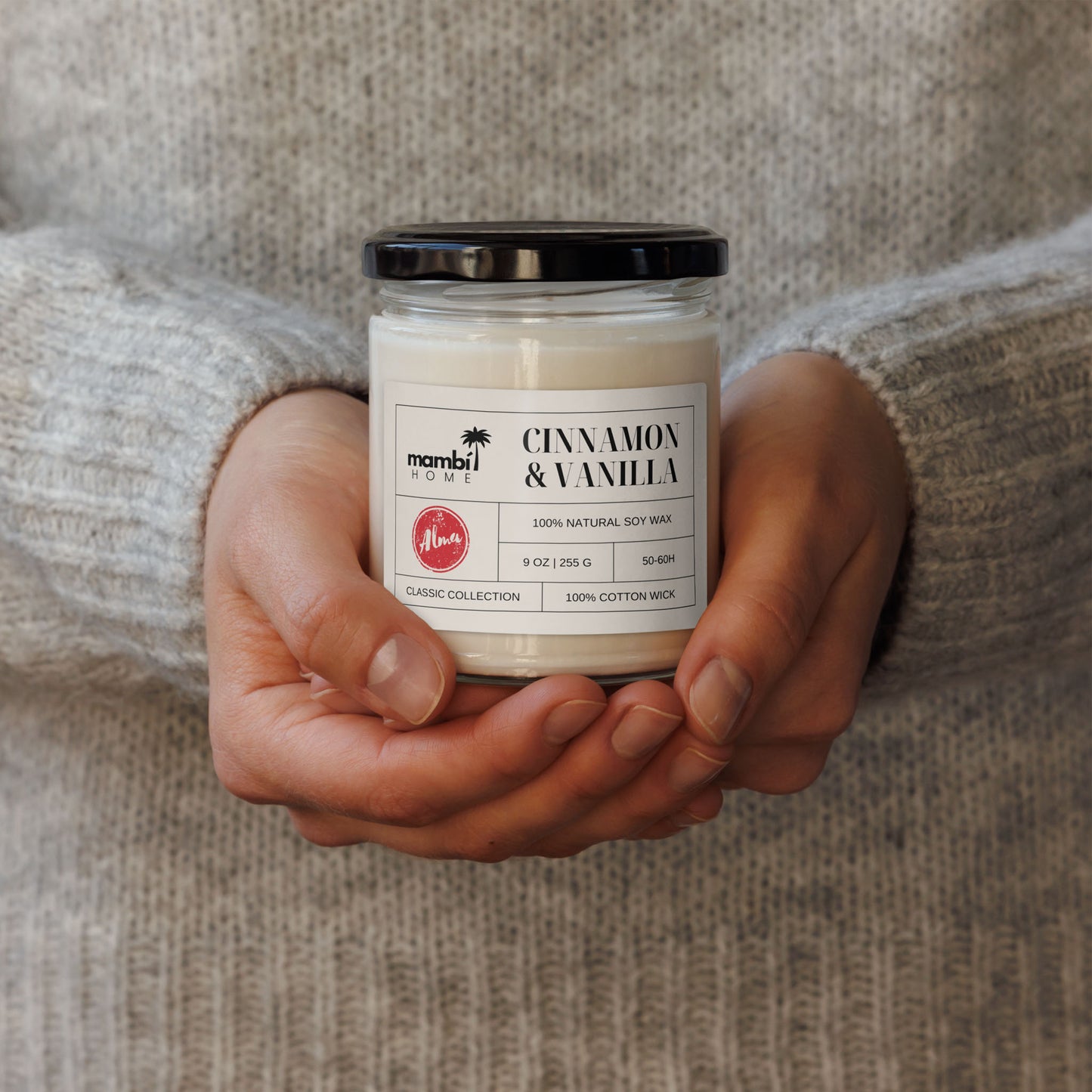 A person wearing a light gray sweater holds a glass jar candle labeled Cinnamon & Vanilla from Mambi Home—an eco-friendly candle with a black lid and white wax.