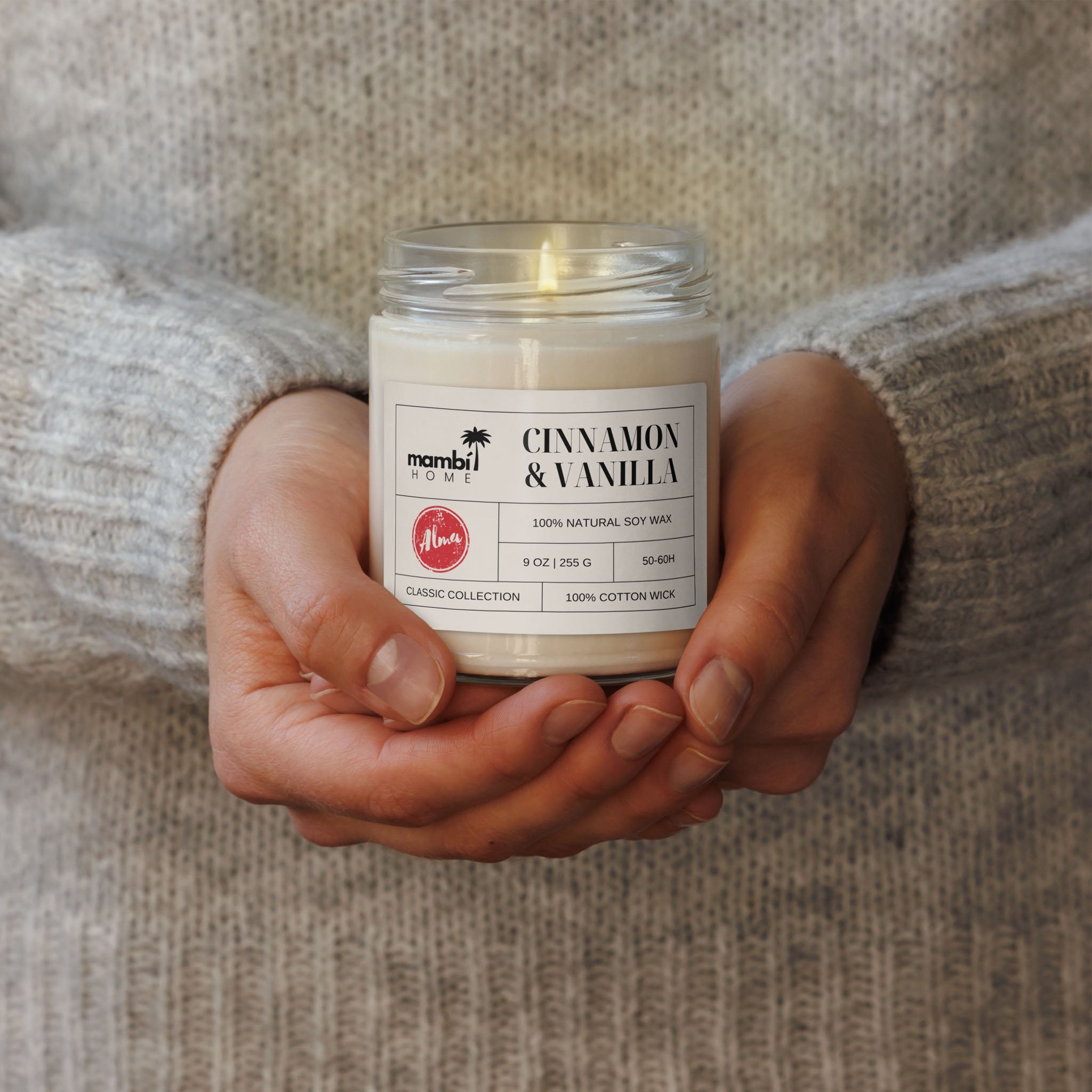 A person in a cozy, light gray sweater holds a lit jar candle labeled Cinnamon & Vanilla with both hands. These scented soy candles are made of 100% natural soy wax for a clean burn and long-lasting fragrance.