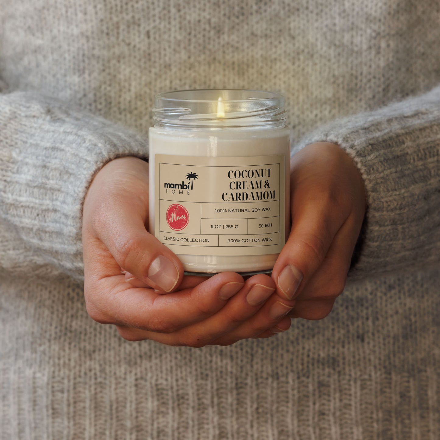 A person in a cozy, light gray sweater holds a glass jar candle labeled Coconut Cream & Cardamom, made from natural soy wax. This Classic Collection piece highlights the appeal of eco-friendly candles for any relaxing space.