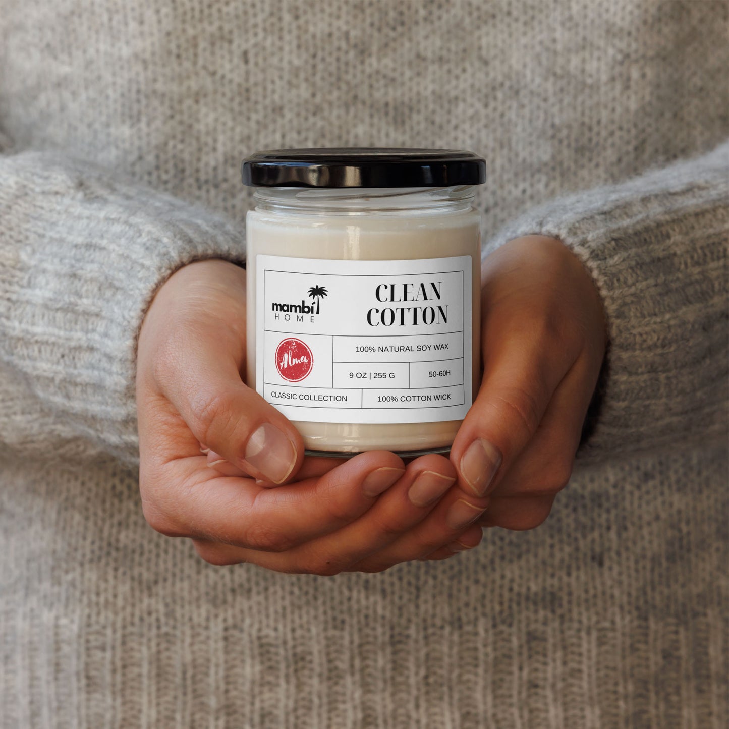 A person wearing a light grey, knit sweater holds a glass jar candle labeled Clean Cotton, made with 100% natural soy wax. This cozy scene highlights the appeal of eco-friendly candles for a fresh and inviting home atmosphere.