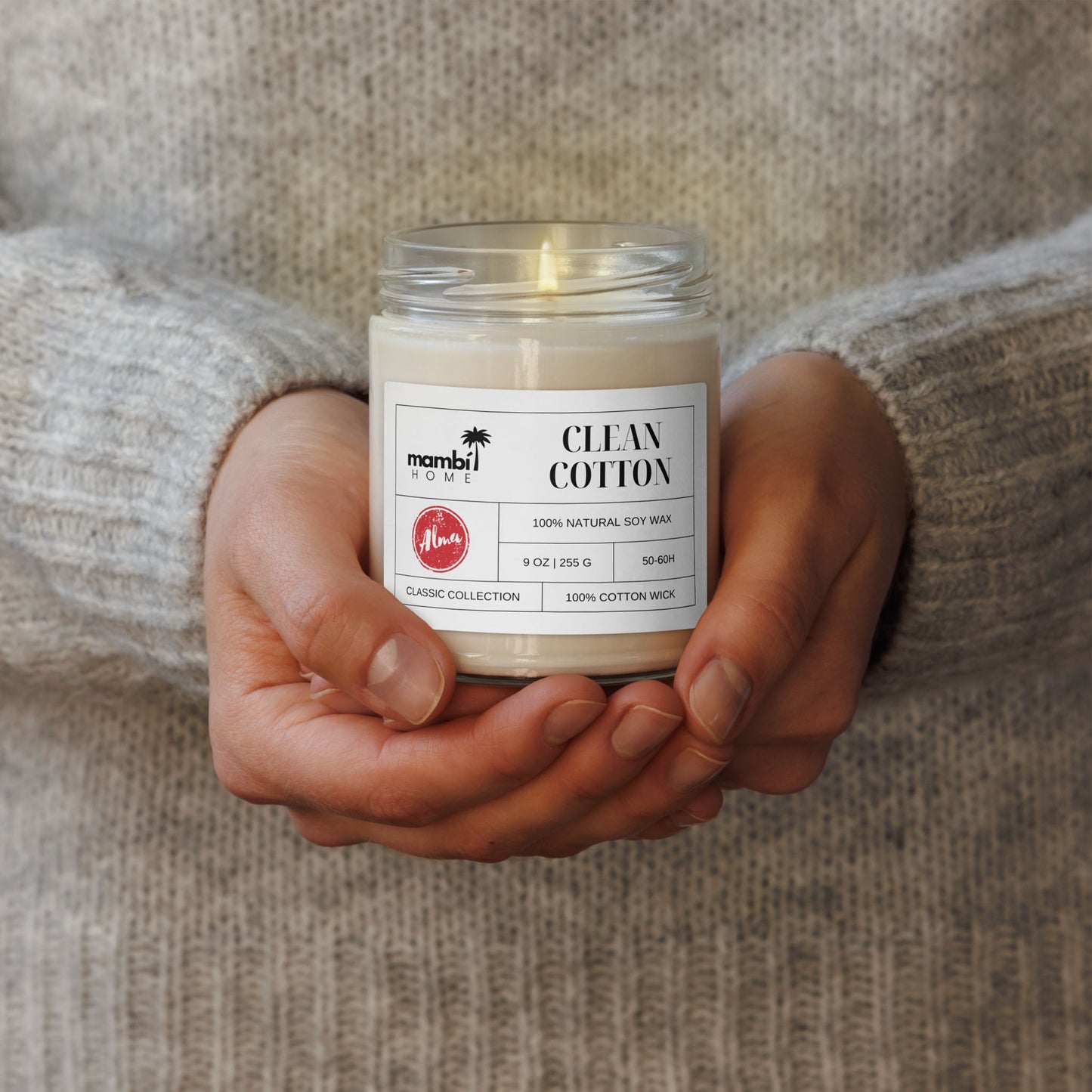 A person wearing a light gray sweater holds a glass jar candle labeled Clean Cotton with both hands. The candle label highlights 100% Natural Soy Wax, showcasing eco-friendly candles from the Classic Collection.