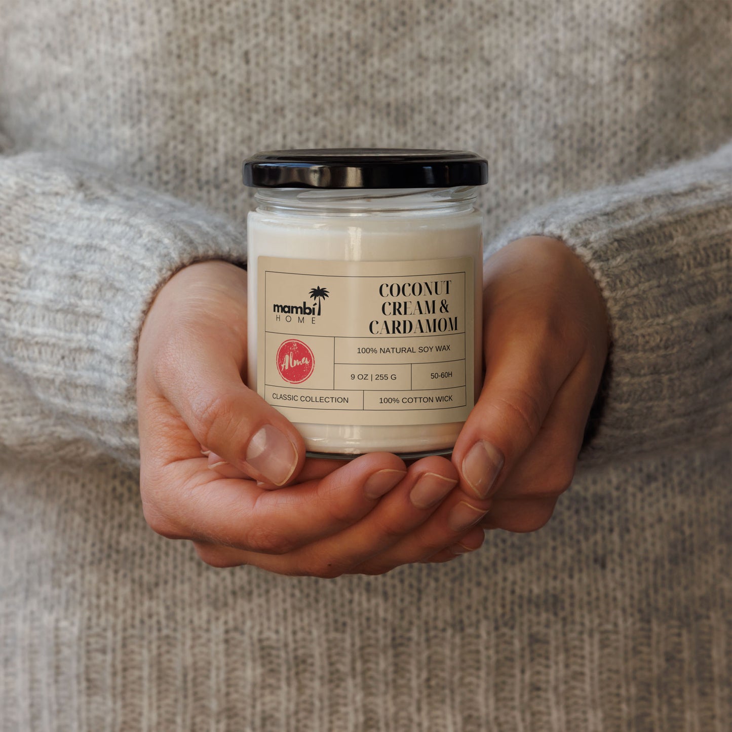 A person wearing a light gray sweater holds a white jar candle labeled Coconut Cream & Cardamom from mambi HOME with both hands. This natural soy wax candle features a black lid, minimalist design, and eco-friendly appeal.