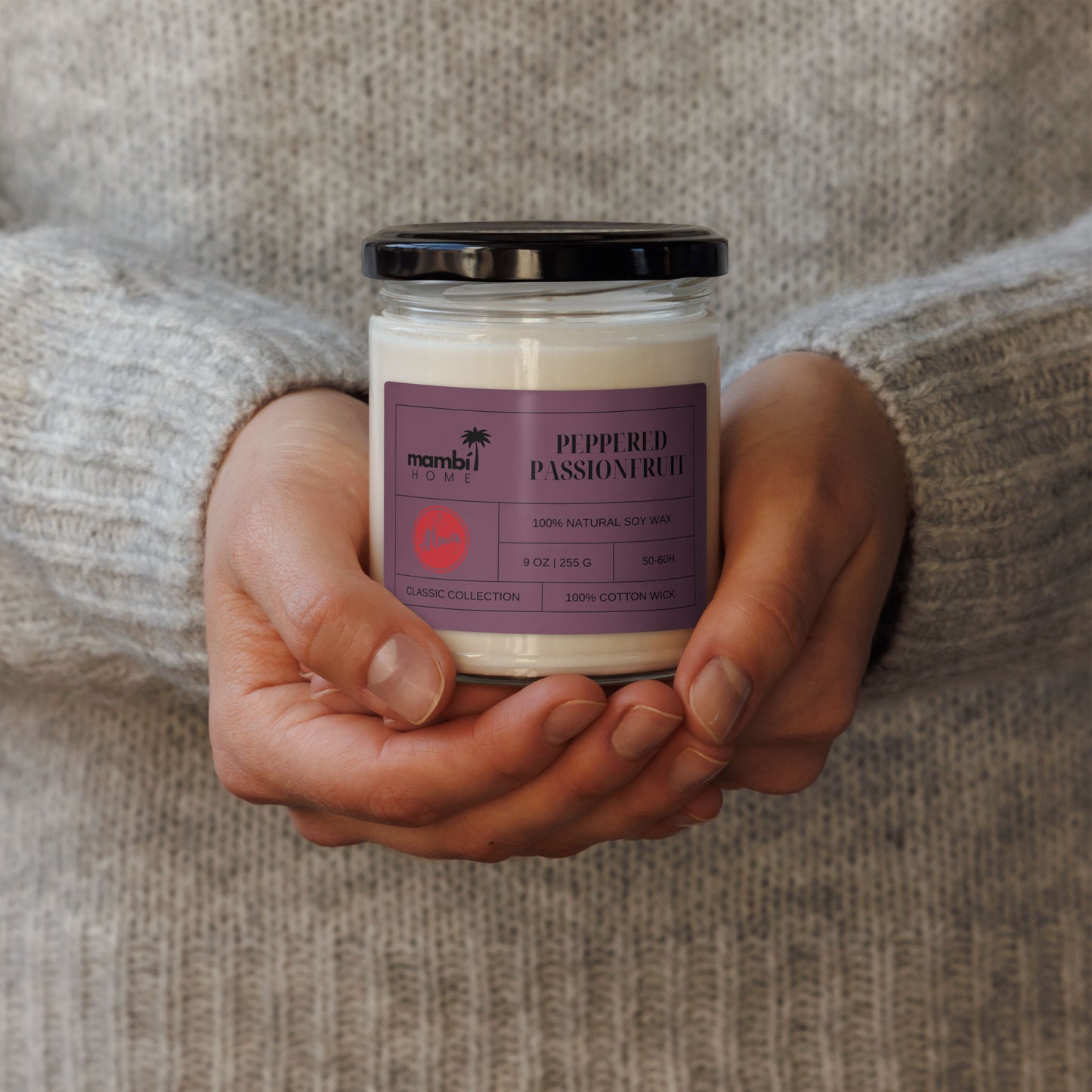 A person in a grey sweater holds a white scented soy candle in a glass jar with a black lid. The label reads Peppered Passionfruit by Mambi Home and describes it as 100% natural soy wax.