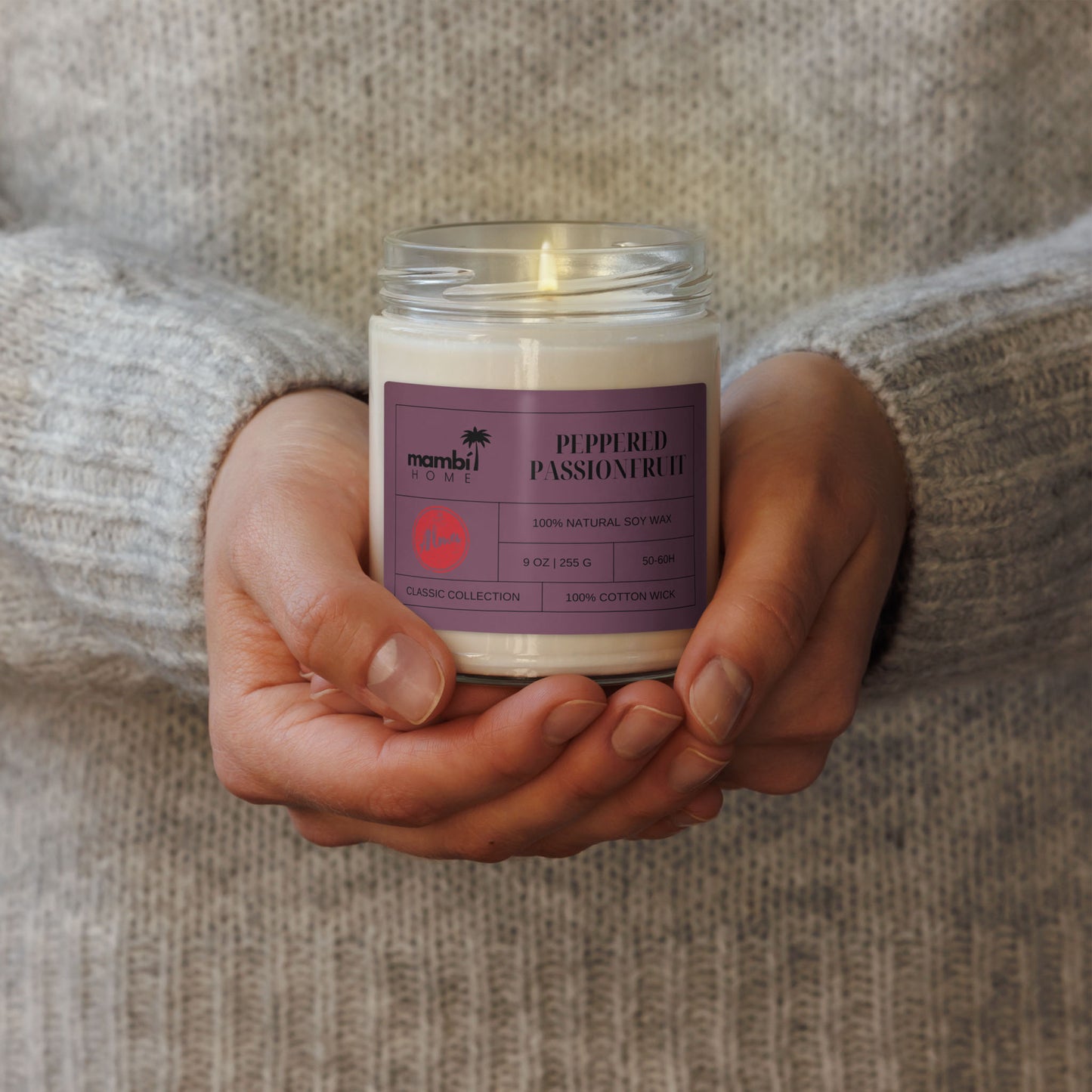 A person wearing a gray knit sweater holds a glass jar candle labeled Peppered Passionfruit by Mambi Home. The label indicates it’s one of their eco-friendly candles made with 100% natural soy wax and a cotton wick.
