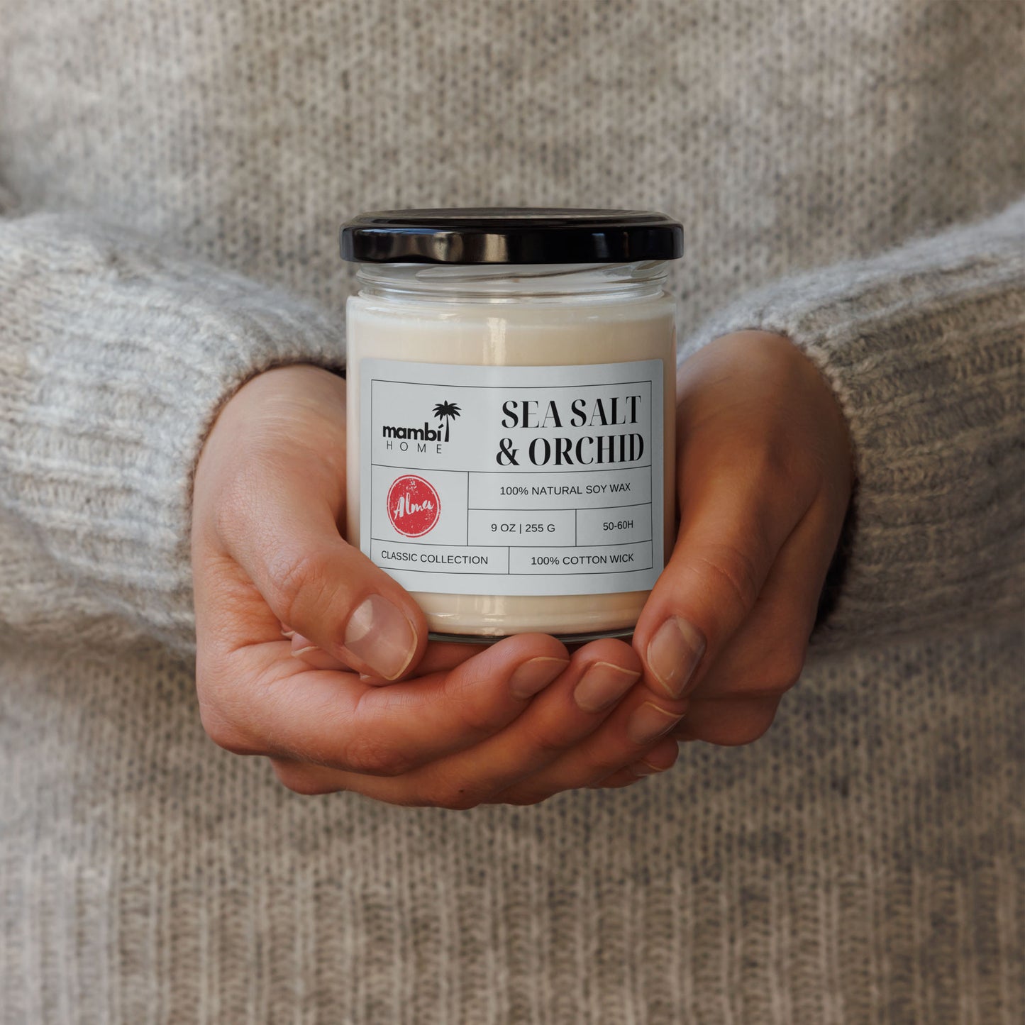 A person in a gray sweater holds a glass jar candle labeled Sea Salt & Orchid from Mambi Home, made with 100% natural soy wax and a cotton wick—perfect for those who love eco-friendly candles.