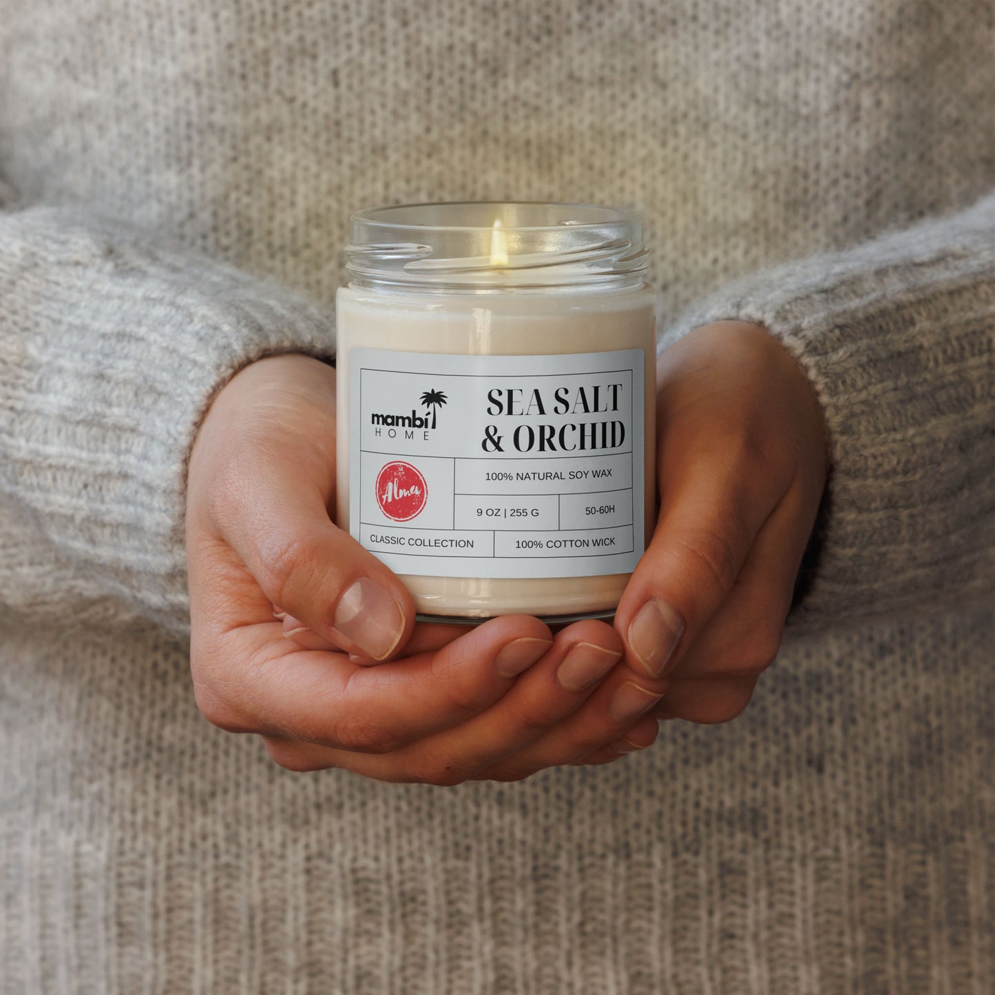 A person in a gray sweater holds a glass jar candle labeled Sea Salt & Orchid from Mambi Home. The label highlights this eco-friendly candle is made with 100% natural soy wax and a cotton wick, part of the classic collection.