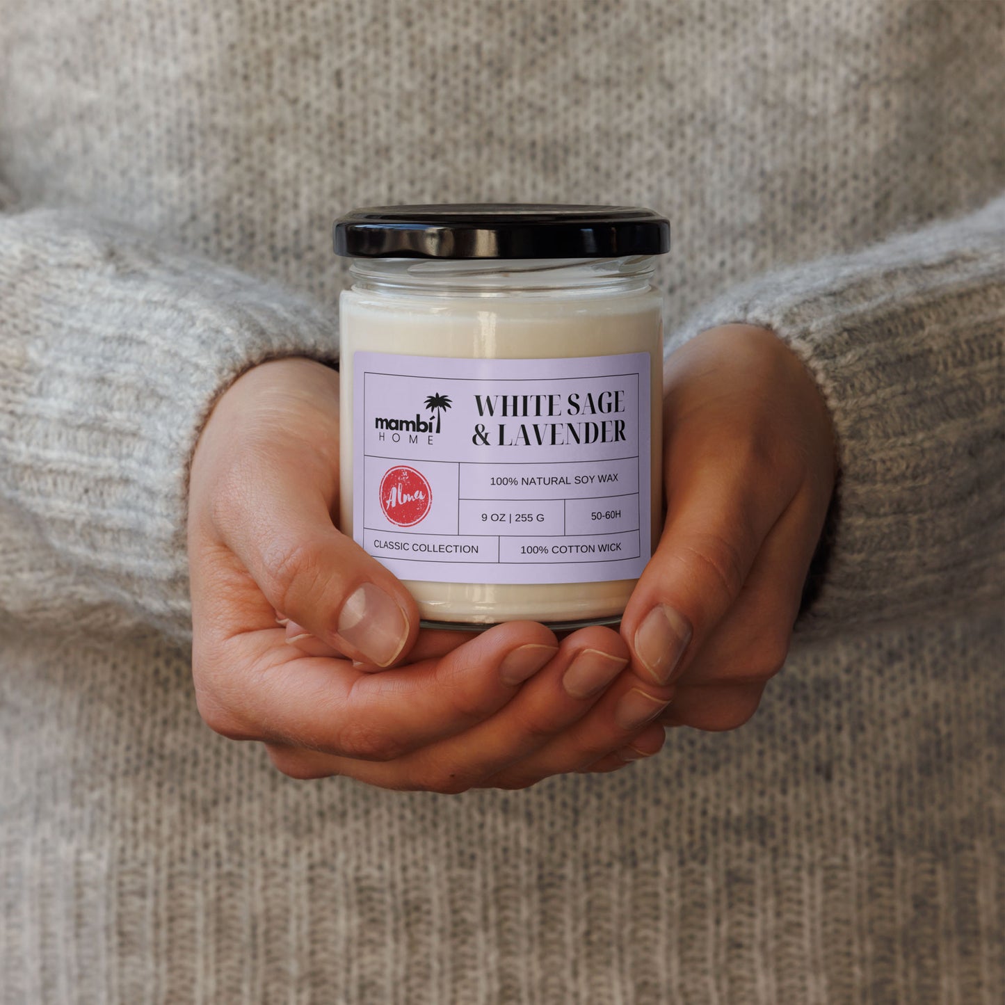 A person in a gray sweater holds a white jar candle labeled White Sage & Lavender by Mambi Home, featuring a black lid and light purple label—one of their eco-friendly scented soy candles.