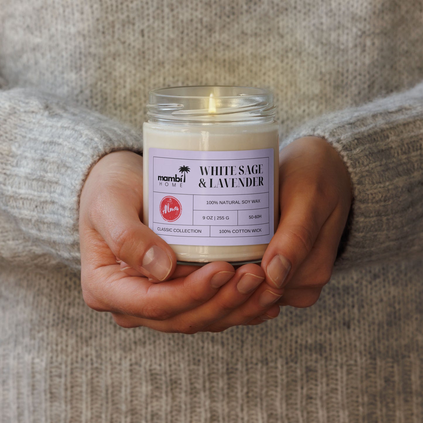 A person in a grey sweater holds a glass jar candle labeled “White Sage & Lavender” from Mambi Home, featuring natural soy wax and a cotton wick—perfect for those who love eco-friendly candles.