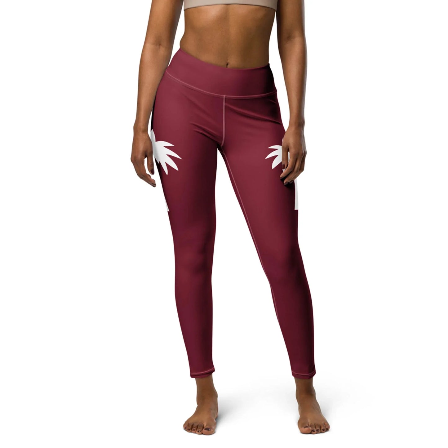 A woman models Printful’s Alpine High-Rise Yoga Leggings in maroon with white palm tree designs, paired with a beige sports bra, embracing athleisure while standing barefoot against a white background.
