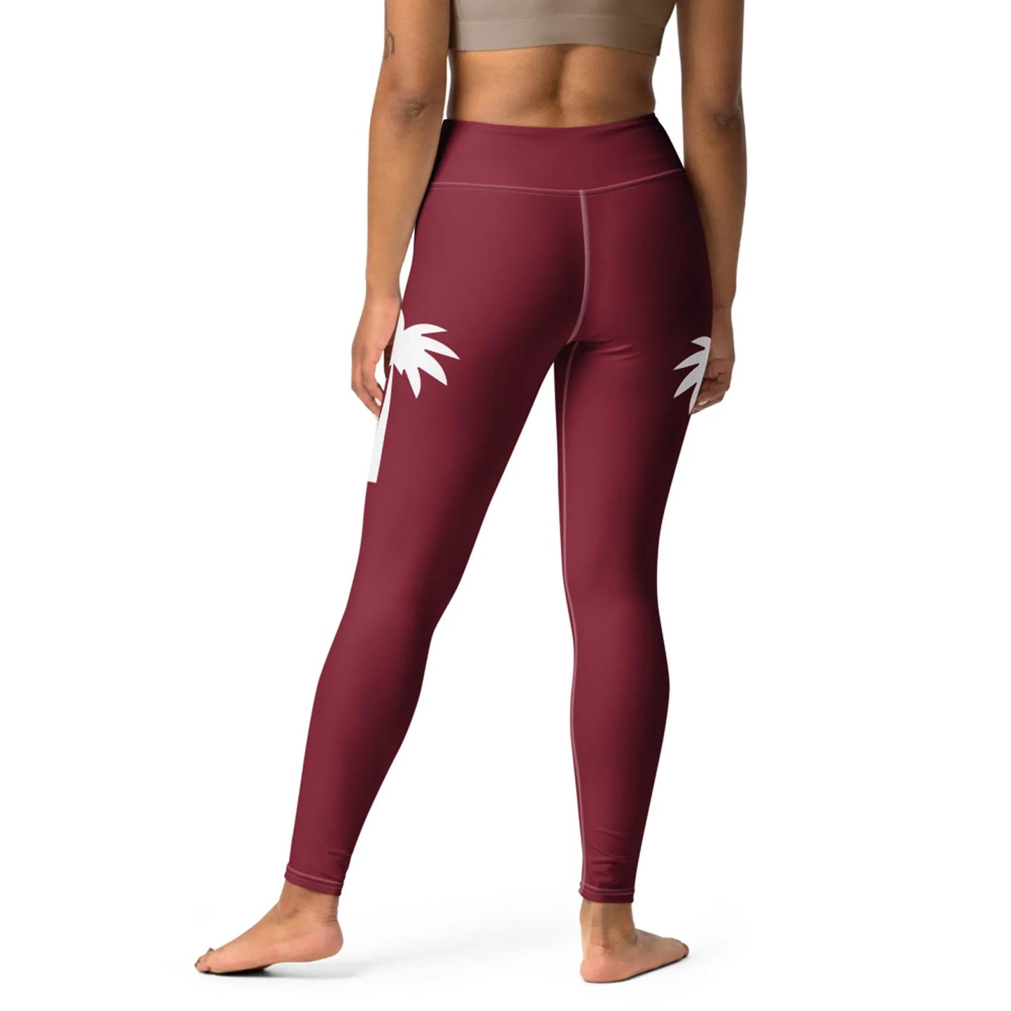 A person stands barefoot, shown from the back, wearing Printfuls Alpine High-Rise Yoga Leggings in maroon with a white palm tree graphic and a beige sports bra—combining comfort and style for any activity.