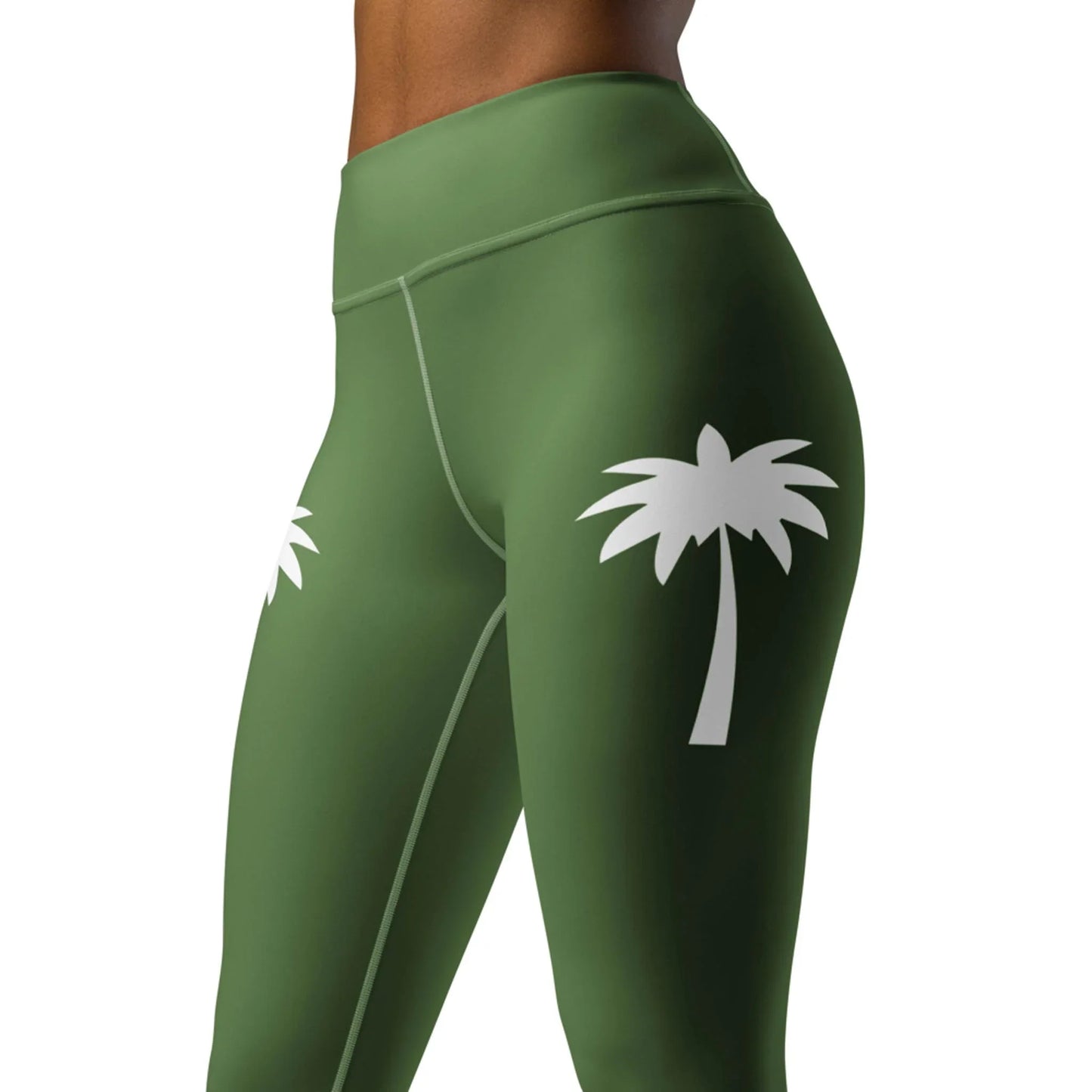A woman models Printfuls Alpine High-Rise Yoga Leggings in olive green, made with four-way stretch fabric and white palm tree graphics on the thighs. The cropped image highlights the torso and upper legs against a plain backdrop.