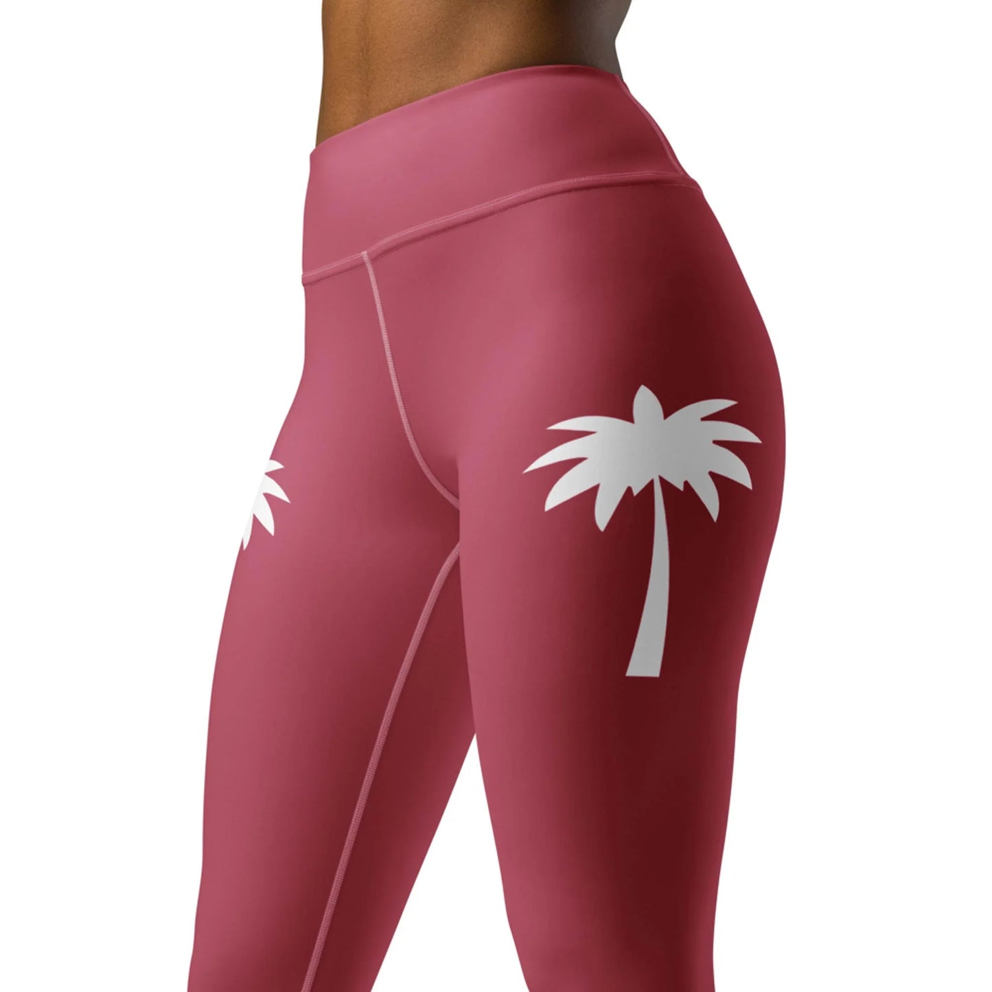 A woman wears Printfuls Alpine High-Rise Yoga Leggings in pink, featuring white palm tree graphics on the hips. The image highlights the lower torso and upper legs, showcasing the athleisure style and chic design of these leggings.