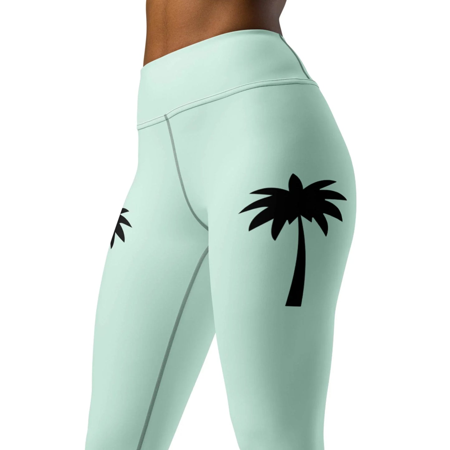 A cropped image shows a person in Printfuls Alpine High-Rise Yoga Leggings for women, featuring light mint green fabric with black palm tree graphics on the outer thighs, highlighting a chic athleisure style.