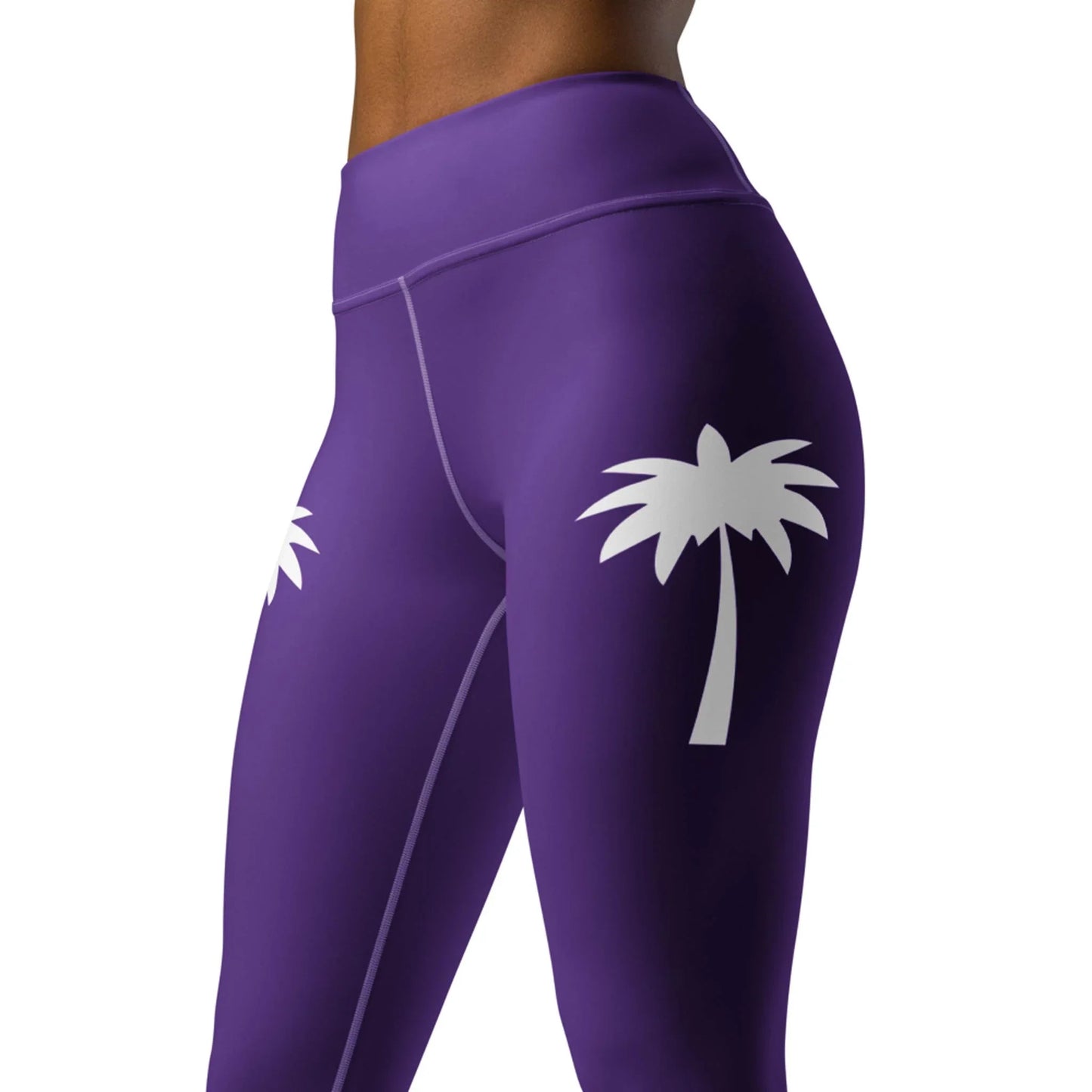 A woman models Printfuls Alpine High-Rise Yoga Leggings in purple, featuring white palm tree graphics on each thigh. The cropped image focuses on the waist and upper legs, showcasing a stylish athleisure look.
