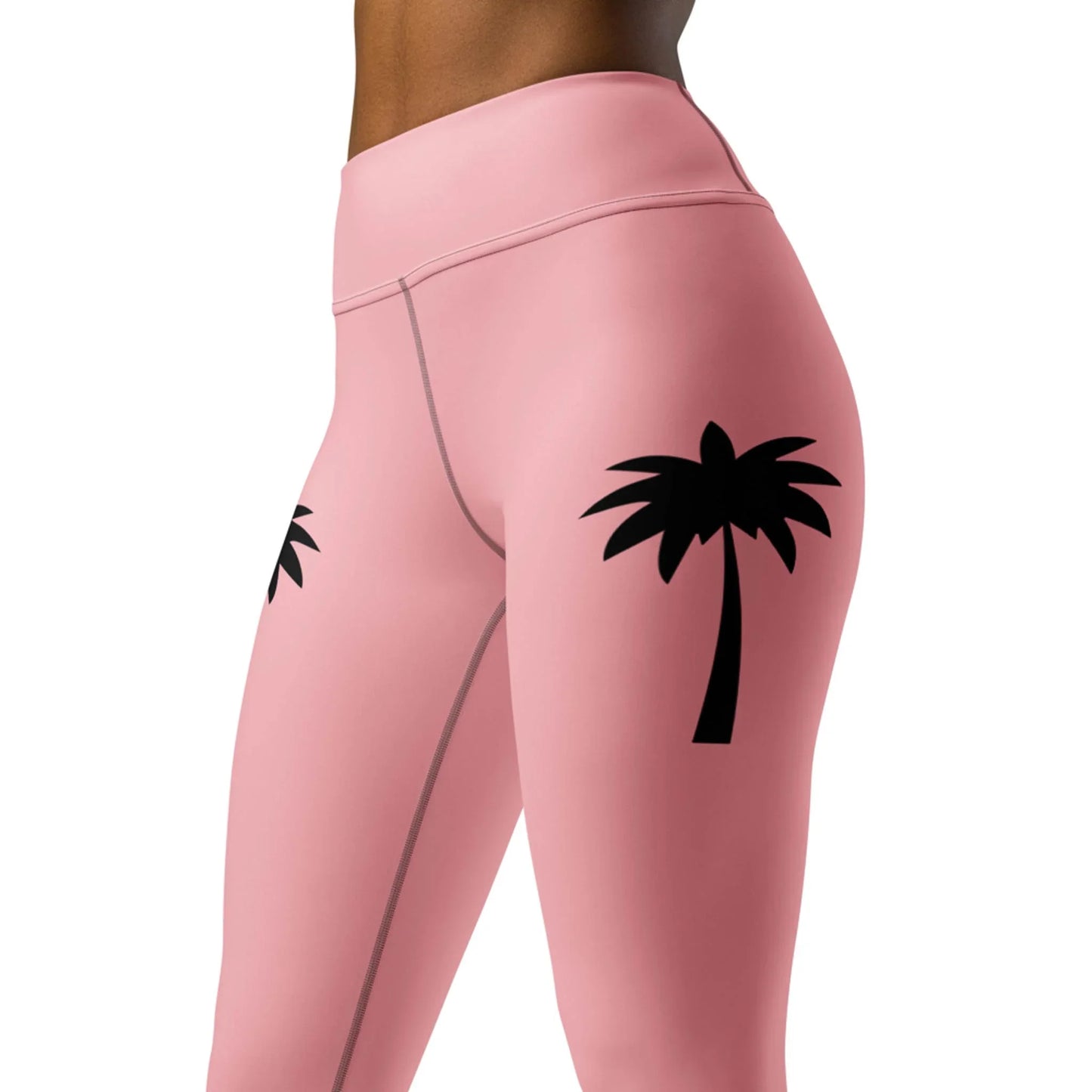 A person wears Printfuls Alpine High-Rise Yoga Leggings for women in pink with black palm tree graphics, shown waist to mid-thigh, showcasing a trendy athleisure style.