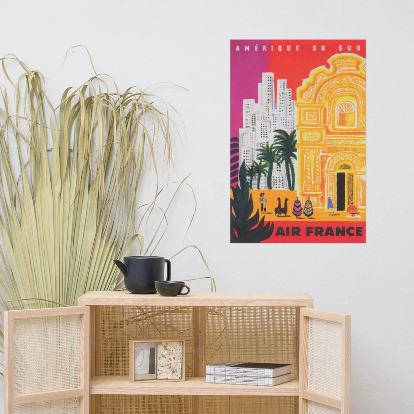 A colorful vintage Air France travel poster for South America, inspired by Mambí style, hangs above a light wooden cabinet with a black teapot and cups. Dried palm leaves sit to the left of the cabinet.