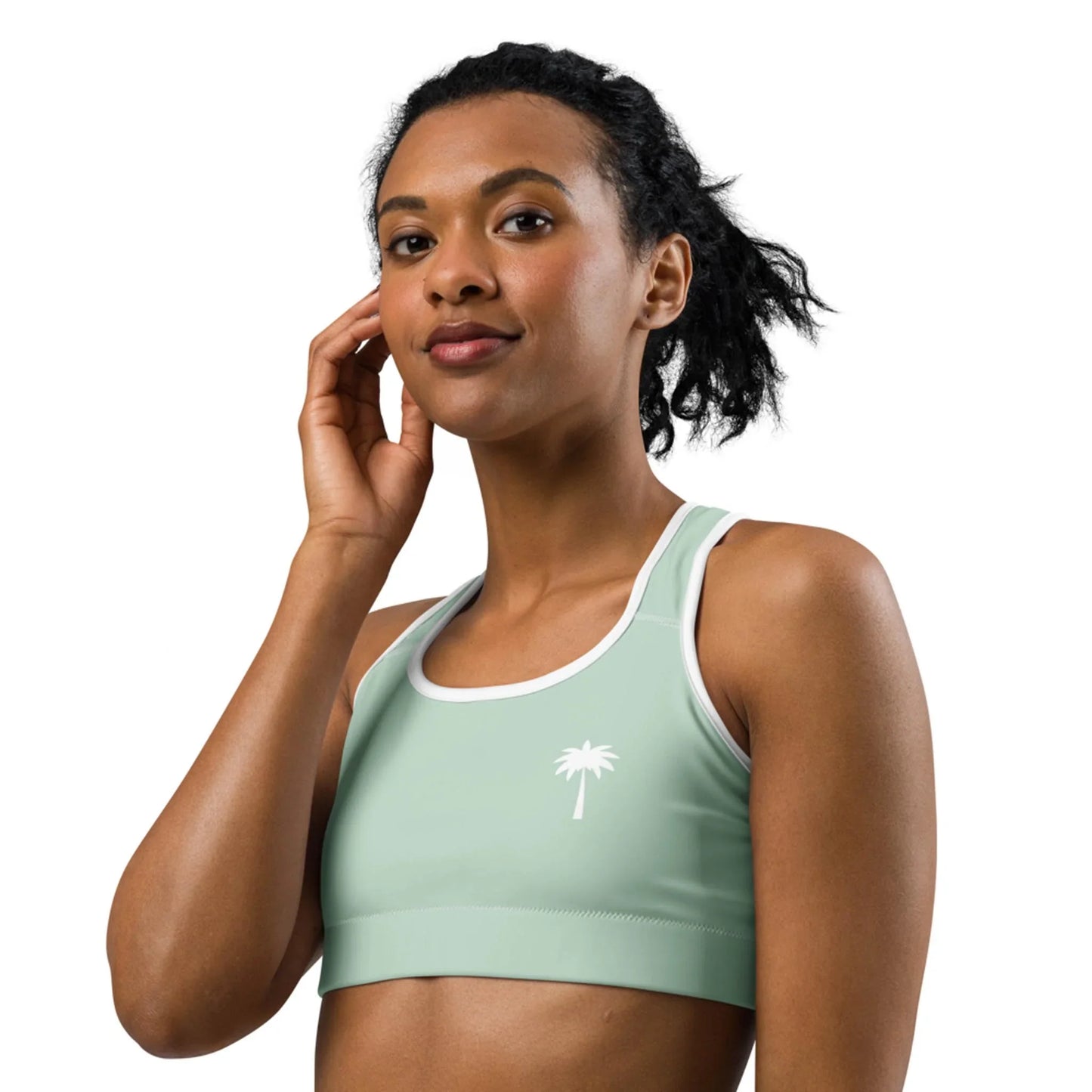 A woman with curly hair in a ponytail wears a light green Mambí athletic crop top with a white palm tree graphic and white trim, posing with one hand touching her face against a plain background.