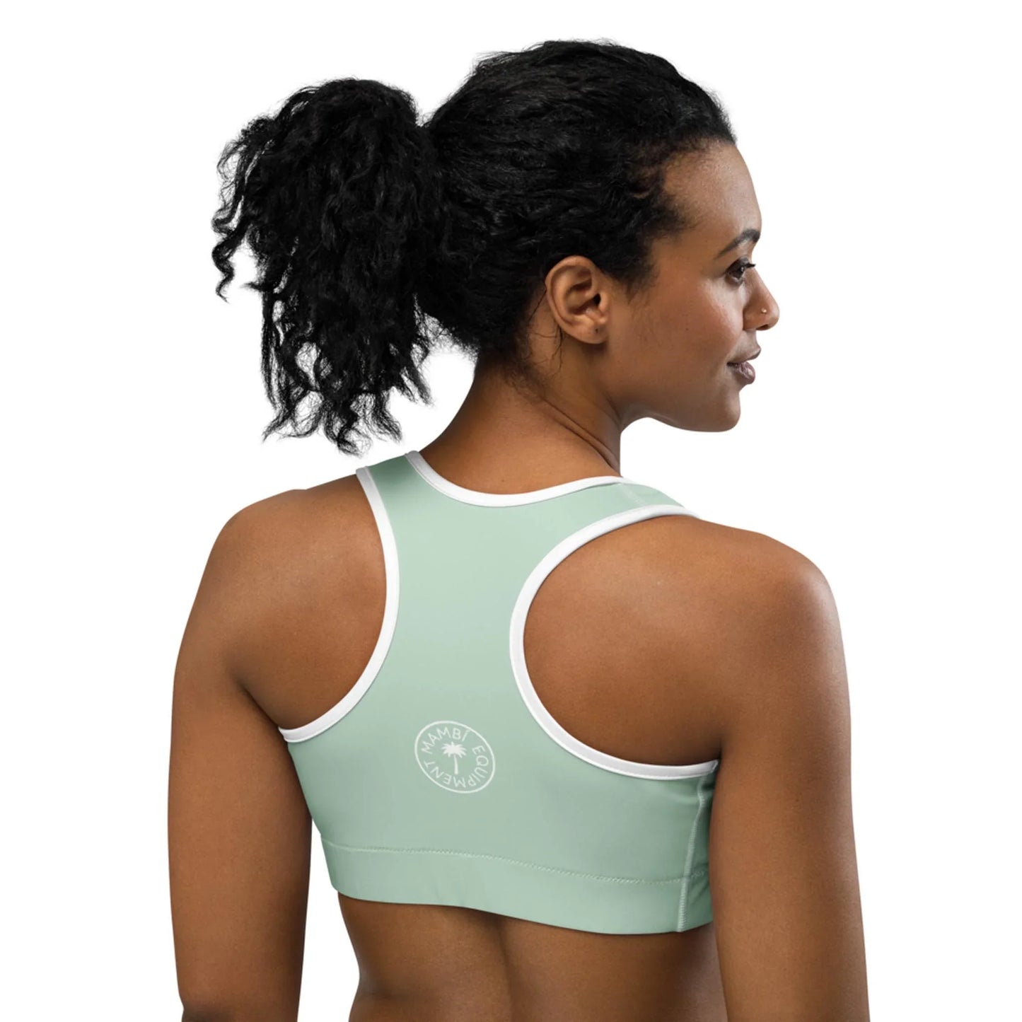 A woman with curly black hair in a ponytail wears a light green Mambí sports bra with white trim. She is facing away, highlighting the back of the bra and its circular white logo on the left side.