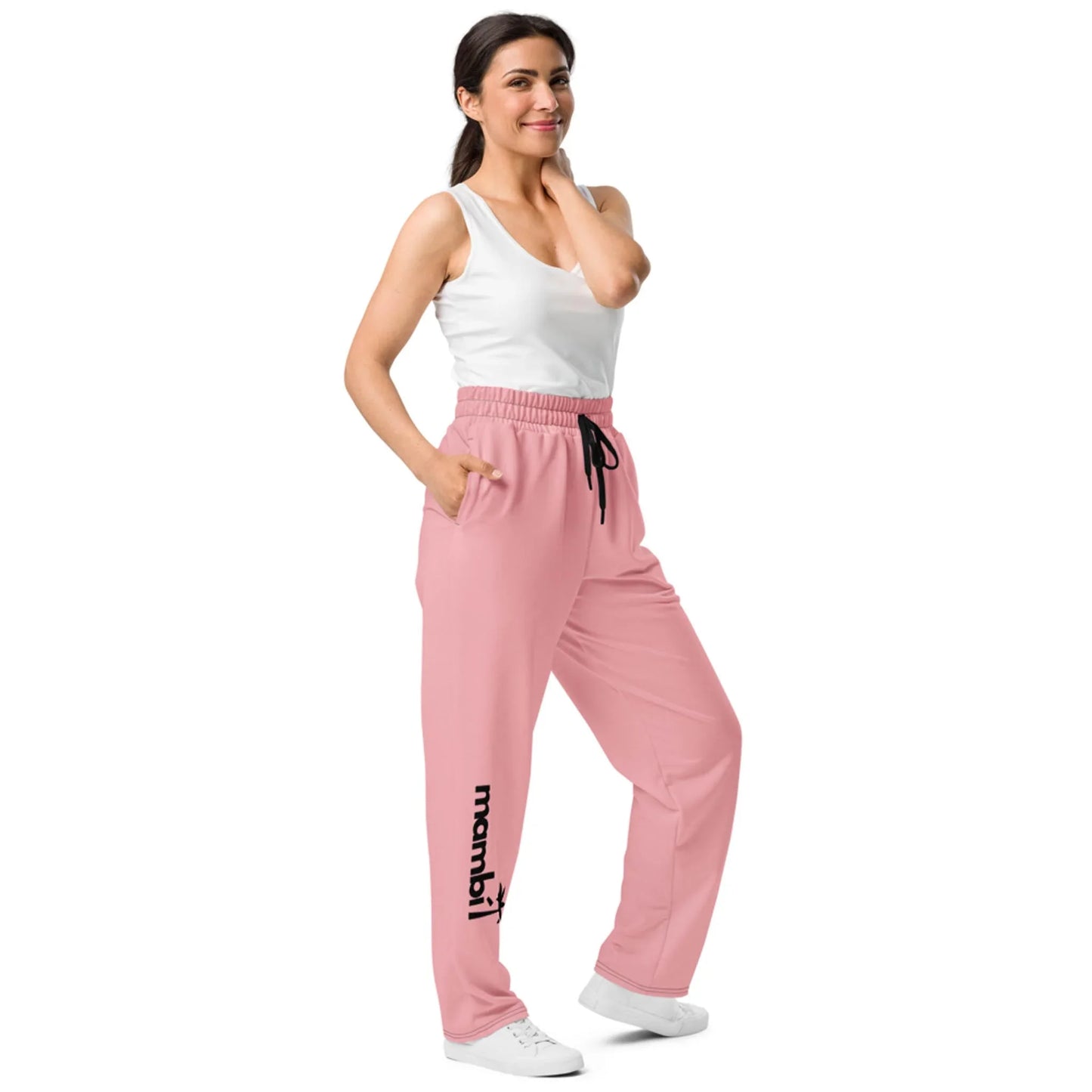 A woman in a white tank top and pink sweatpants with Mambí near the left ankle stands smiling, one hand on her neck and the other in her pocket. She is wearing white sneakers.