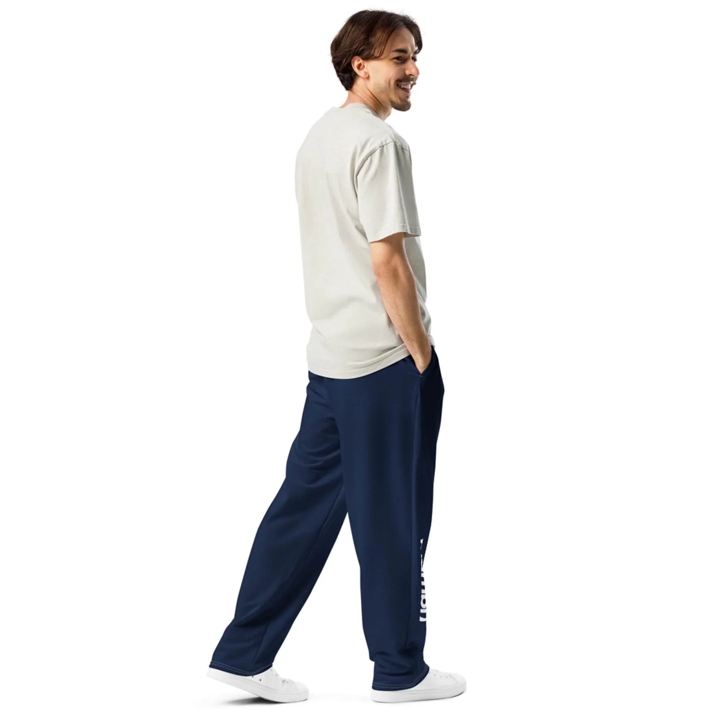 A man stands smiling, facing sideways and looking back. He wears a light gray Mambí t-shirt, navy blue sweatpants with white text on the leg, and white sneakers. His hands are in his pants pockets against a plain white background.