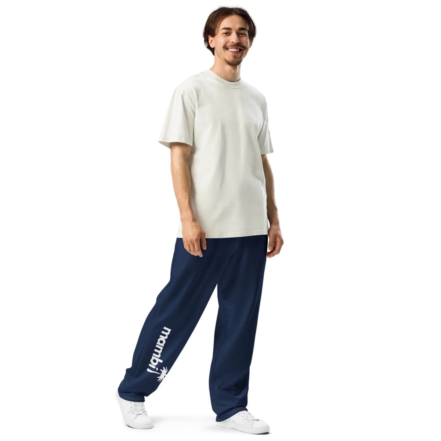 A smiling man stands wearing a cream-colored t-shirt, navy blue Mambí pants with a palm graphic on the left leg, and white sneakers against a white background.