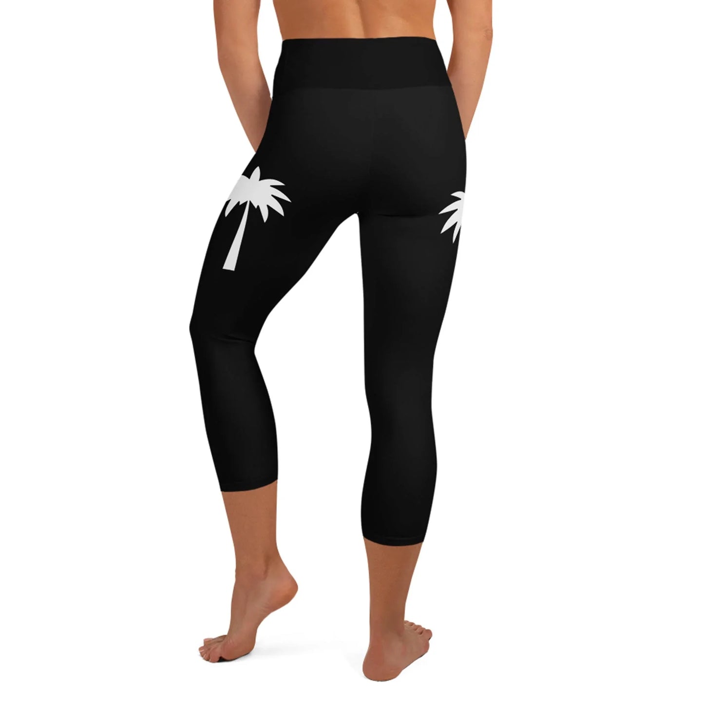 A woman models the Printful Aventura High-Rise Yoga Capri Leggings, featuring white palm tree graphics on the back hips. The four-way stretch yoga capris are shown barefoot from behind against a white background.
