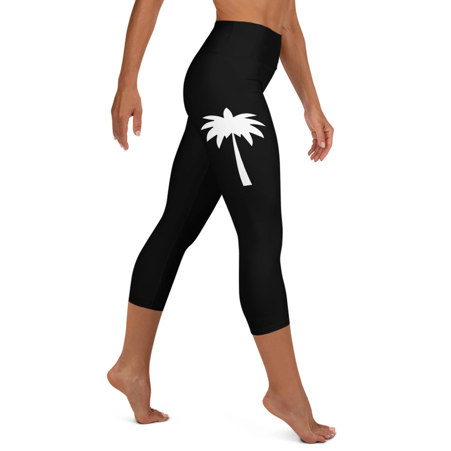 A woman models Printfuls Aventura High-Rise Yoga Capri Leggings for women, featuring a white palm tree graphic on the upper side. Made with four-way stretch fabric, shes barefoot against a plain white background.