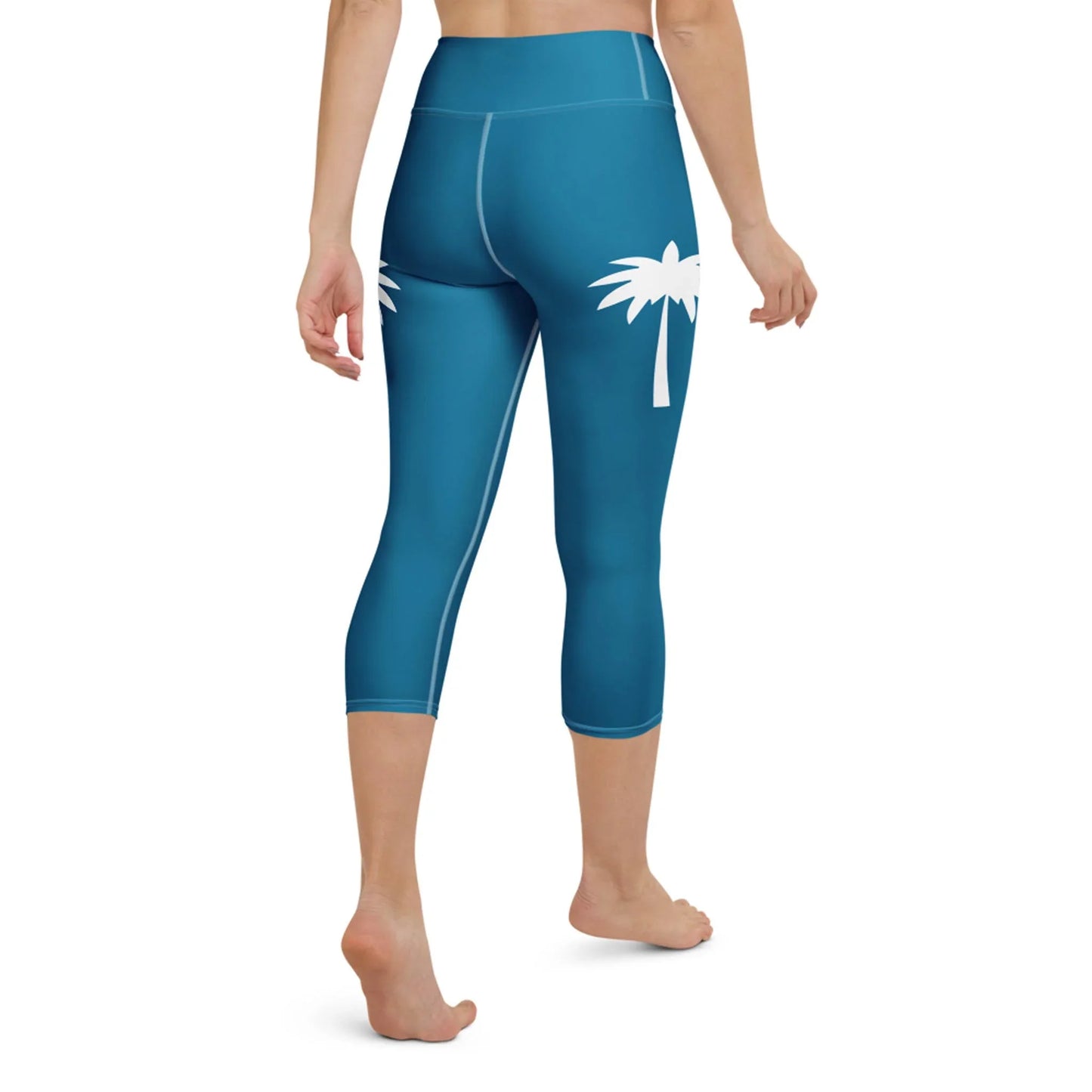A woman models the Printful Aventura High-Rise Yoga Capri Leggings for women in teal, featuring a white palm tree graphic on the left hip. She stands barefoot, back view, against a plain white background. Part of our activewear collection.