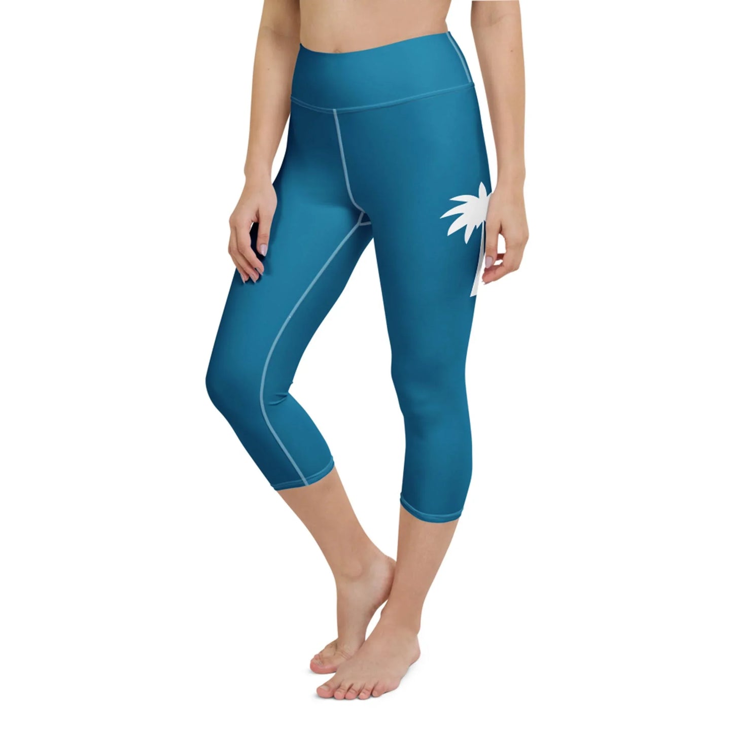 A woman stands barefoot against a white background wearing Printful Aventura High-Rise Yoga Capri Leggings in teal, featuring white seams, a left hip palm tree graphic, and four-way stretch fabric.