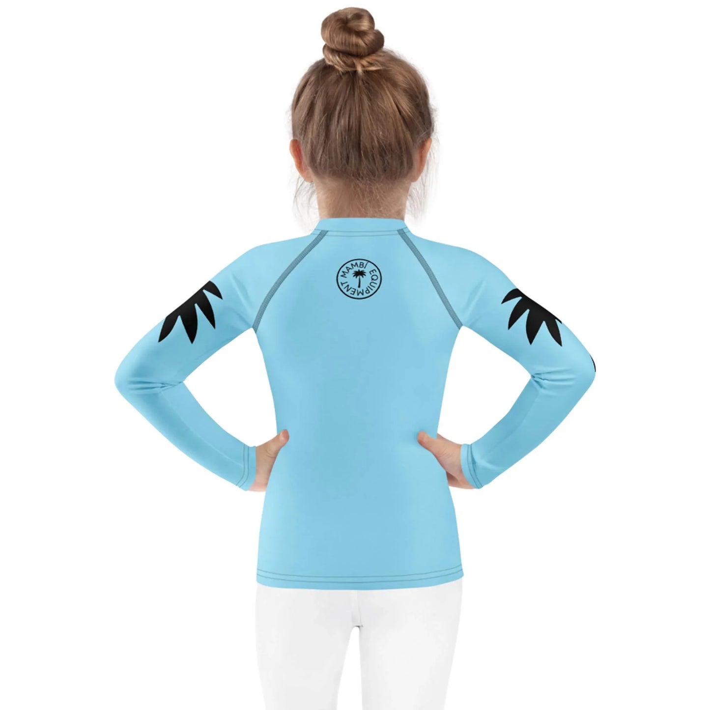 A young child with light brown hair in a bun faces away, wearing a light blue long-sleeve Mambí rash guard with black palm designs on the shoulders and a circular logo on the upper back. The child is also wearing white pants.
