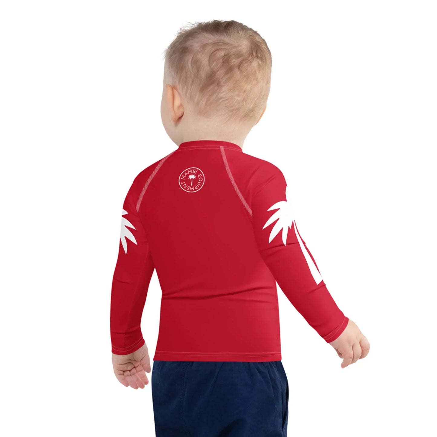 A toddler with short blonde hair, seen from behind, wears a Mambí red long-sleeve rash guard with white palm prints on the shoulders and dark blue pants, standing against a plain white background.