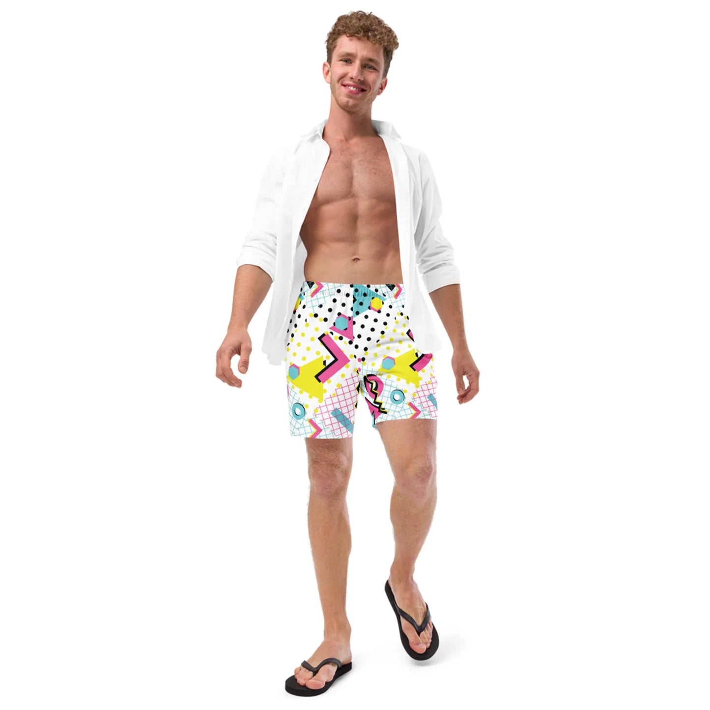 A smiling man in a white open shirt, colorful Mambí patterned shorts, and black flip-flops stands against a white background, looking relaxed and confident.