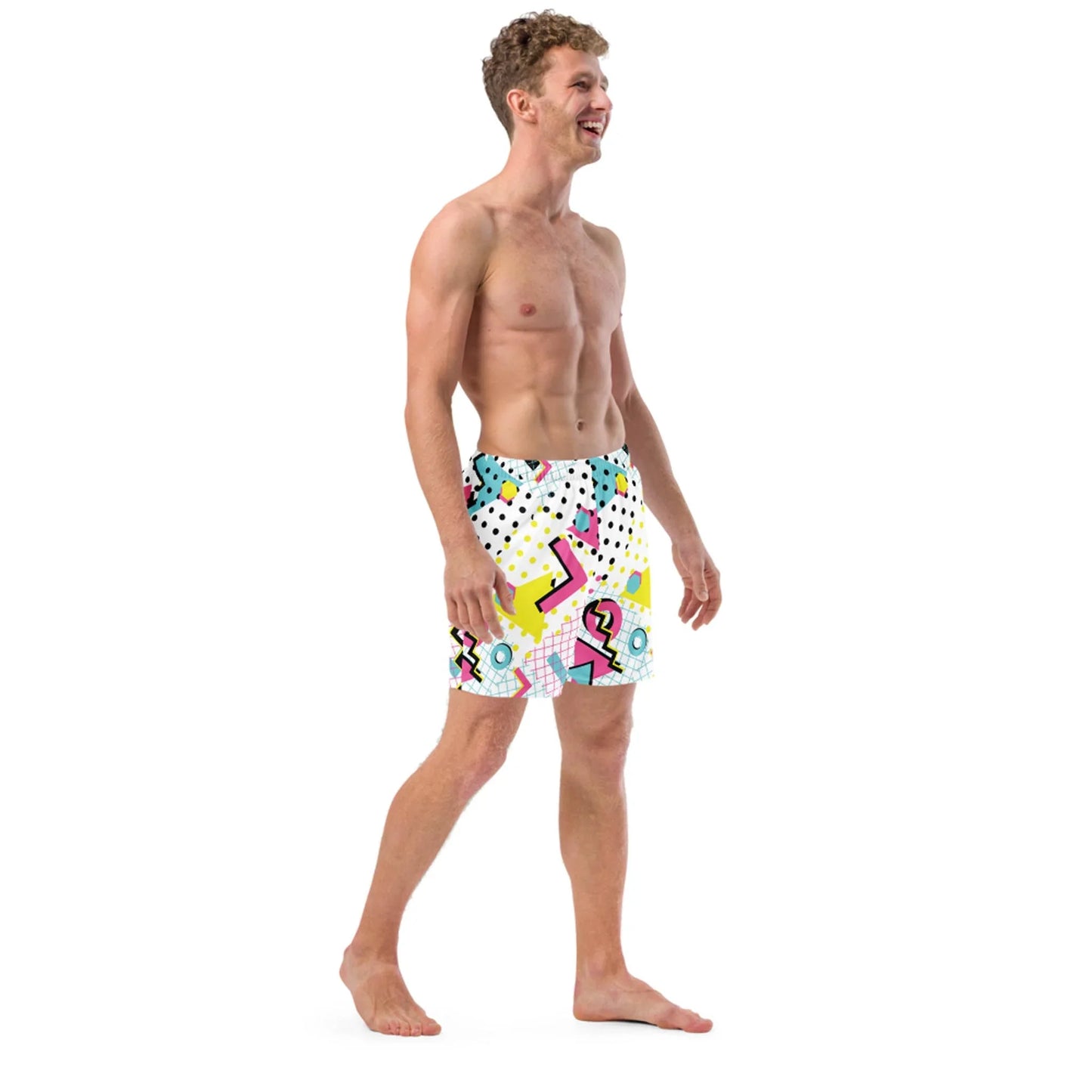 A smiling man stands barefoot, wearing colorful Mambí shorts with a bright geometric and abstract pattern featuring yellow, pink, blue, and black shapes on a white background.