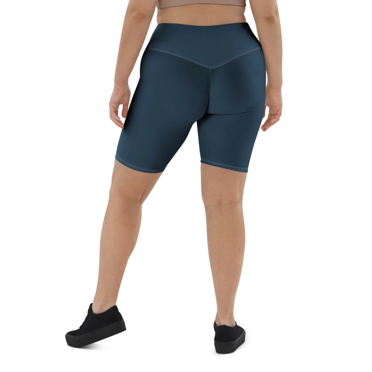 A person stands facing away, wearing a beige Mambí crop top, high-waisted dark blue biker shorts, and black sneakers against a plain white background.
