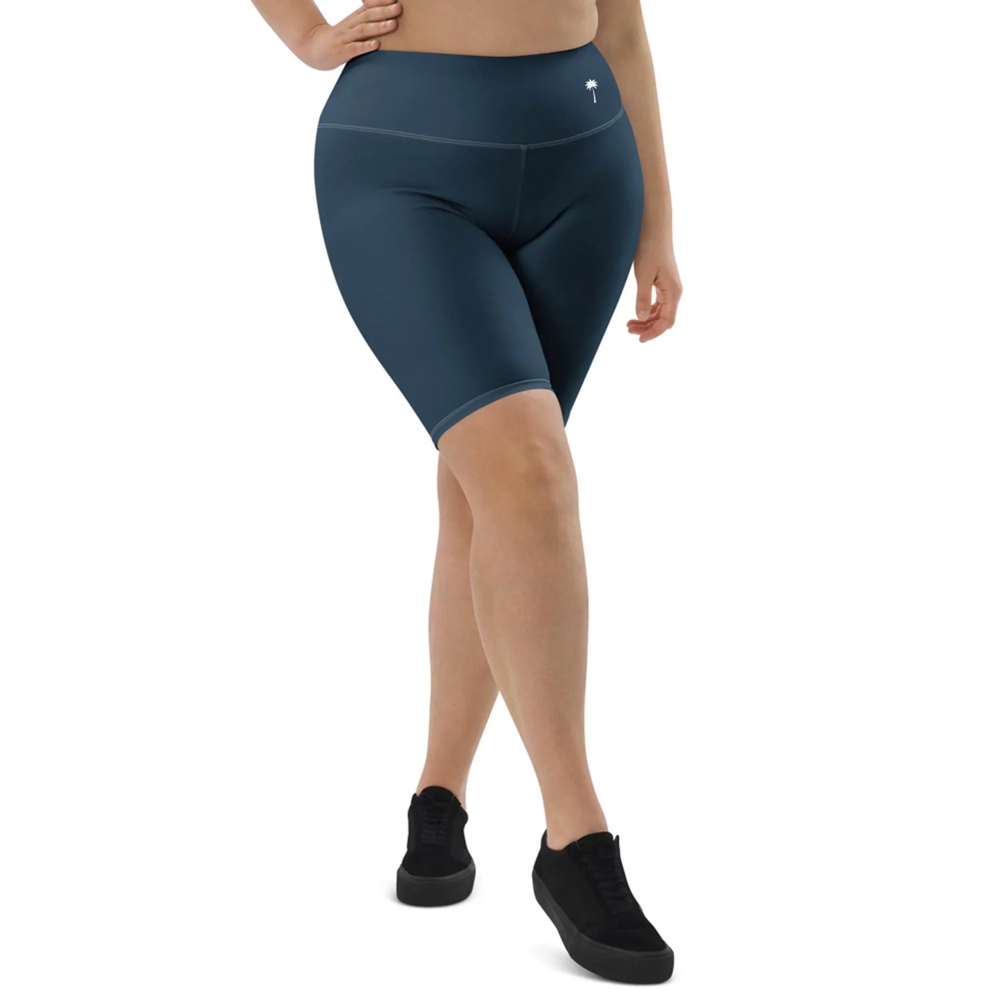 A person wearing high-waisted, dark blue Mambí bike shorts and black sneakers stands with one leg slightly forward, cropped from the shoulders down against a white background.