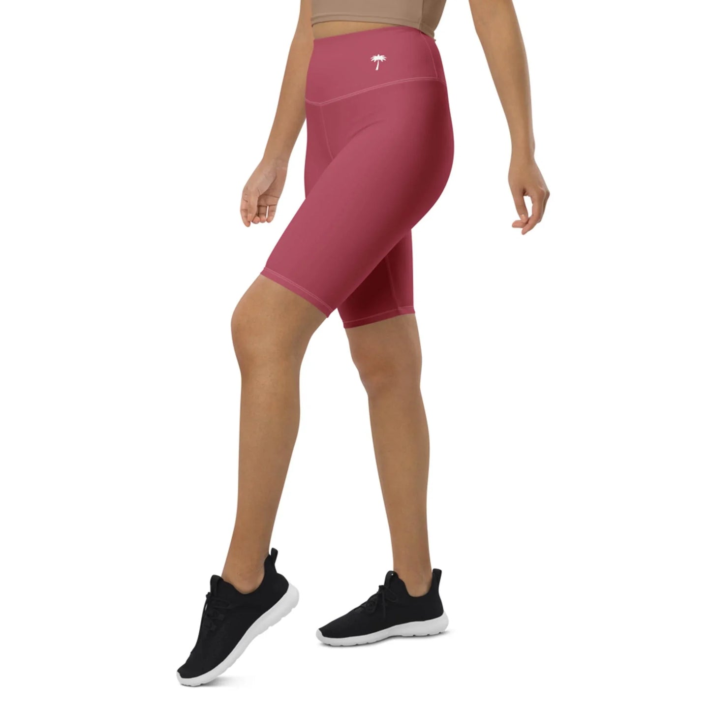 A person wearing high-waisted, dark pink Mambí bike shorts, a cropped beige top, and black athletic shoes stands against a white background. The person’s head is not visible in the image.