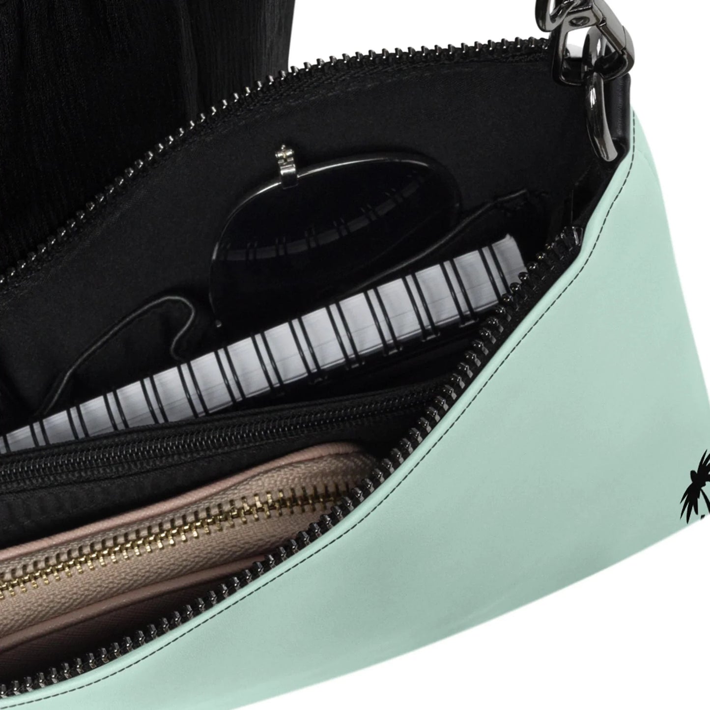 A close-up of the open mint-green Blake Crossbody Bag by Printful with adjustable straps shows a spiral notebook, black sunglasses, and a beige zippered pouch neatly organized inside.