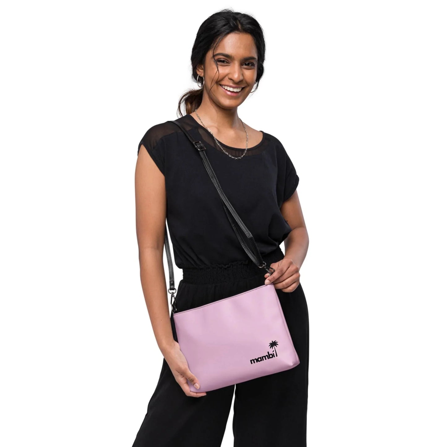 A smiling woman in black holds the Printful Blake Crossbody Bag, featuring a mambi logo and palm tree graphic on premium faux leather with adjustable straps. She stands against a plain white background.
