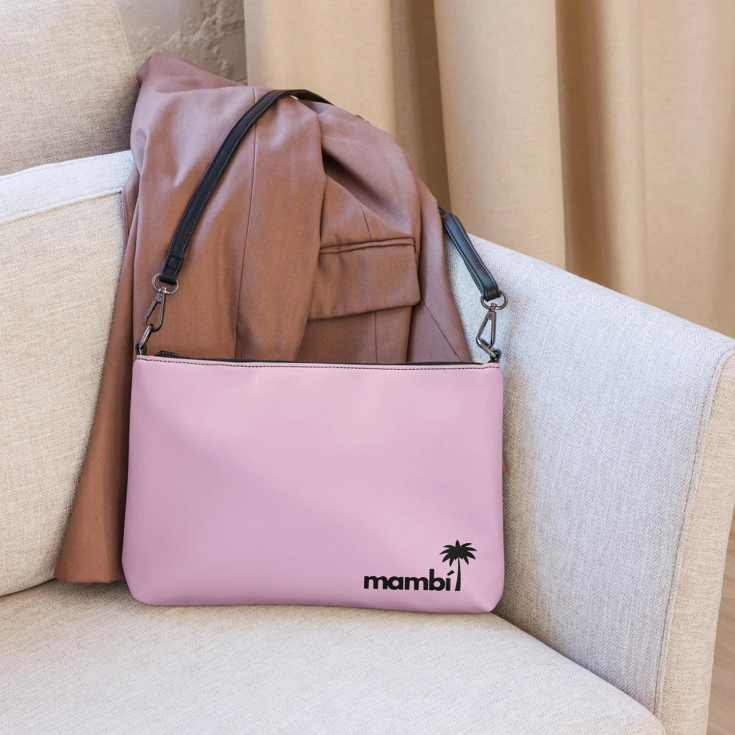 The Blake Crossbody Bag by Printful, crafted in light pink premium faux leather with black adjustable straps and a black palm tree mambi logo, rests on a beige sofa beside a brown blazer.