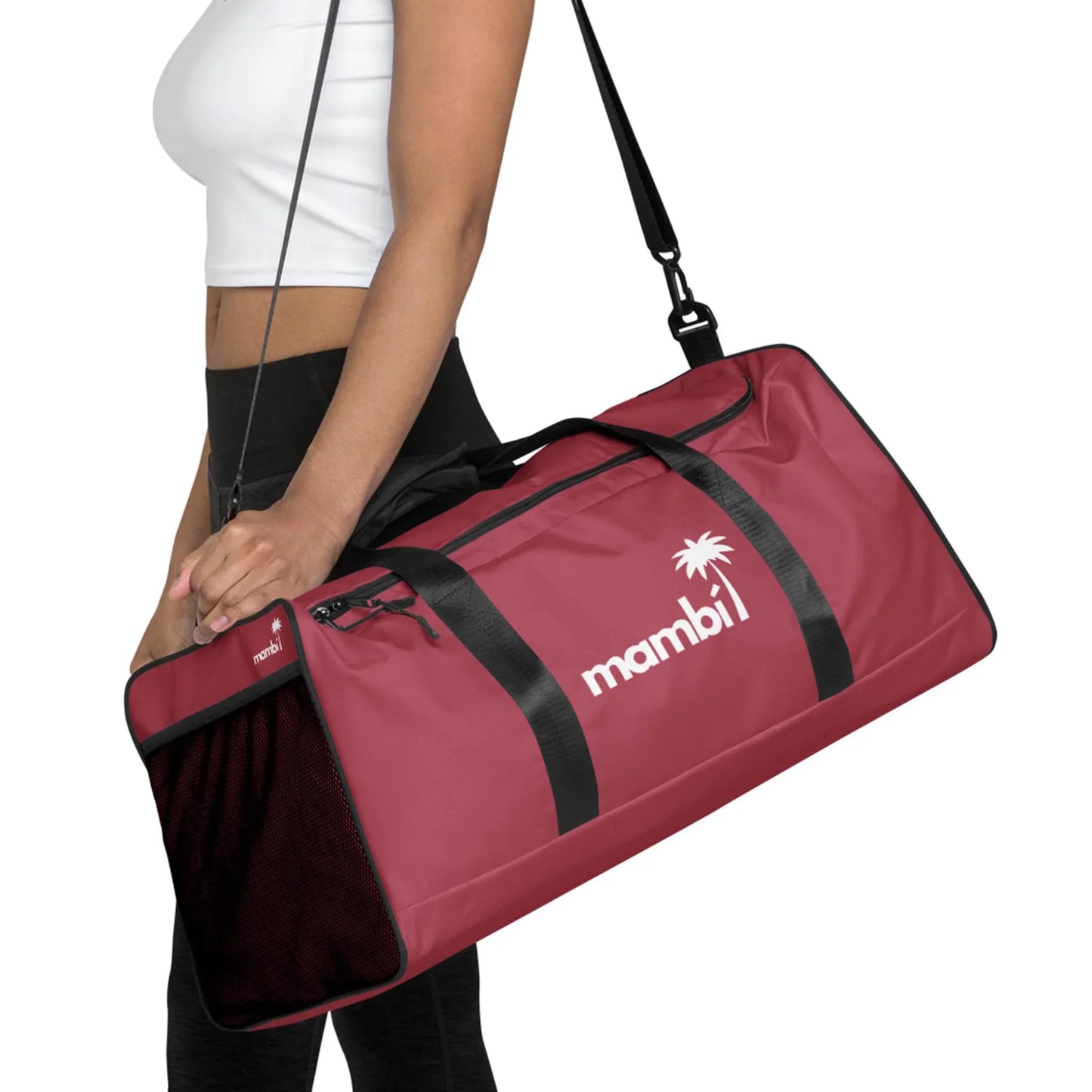 A person wearing a white crop top and black leggings carries a large maroon Mambí duffel bag with black straps and a palm tree logo printed in white.