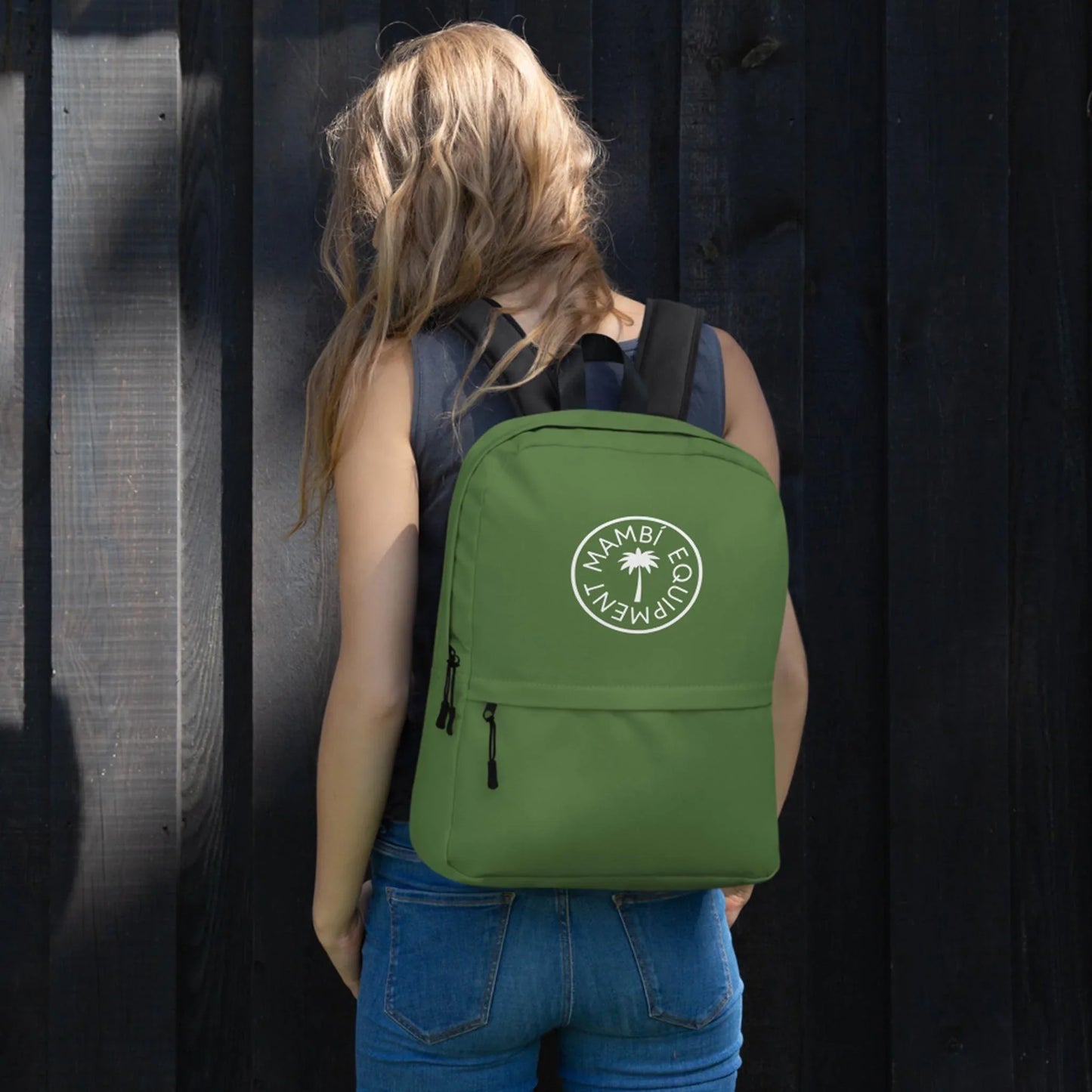 A woman with long, wavy blonde hair, dressed in a black sleeveless top and blue jeans, stands facing a dark wooden fence. She carries the Printful Boulevard East All-Purpose Backpack featuring a circular palm tree logo and hidden back pocket.