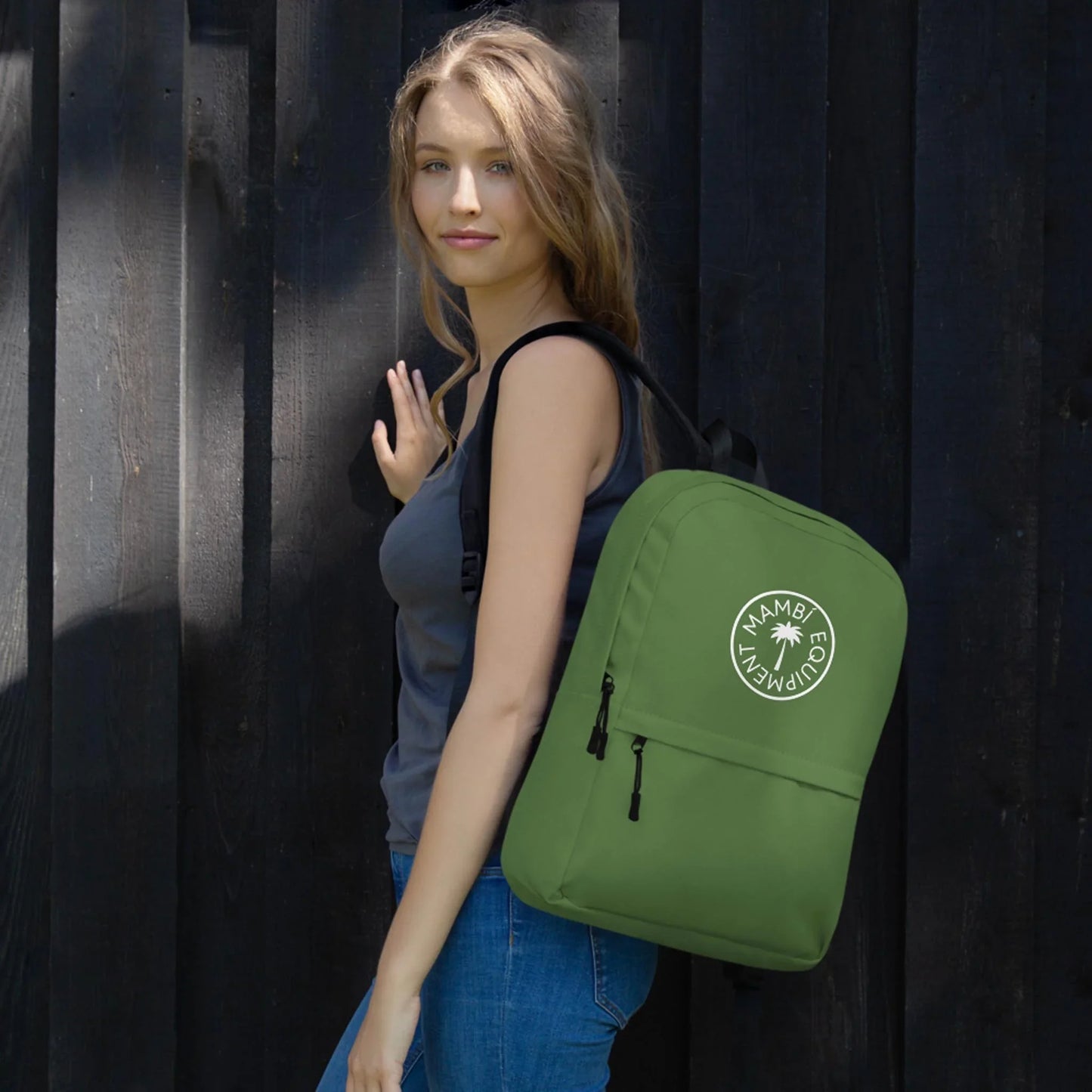A young woman with long, light brown hair wearing a dark tank top and jeans stands against a dark wooden wall, carrying the Boulevard East All-Purpose Backpack by Printful with a circular MAMBO ECO ADVENTURE logo.
