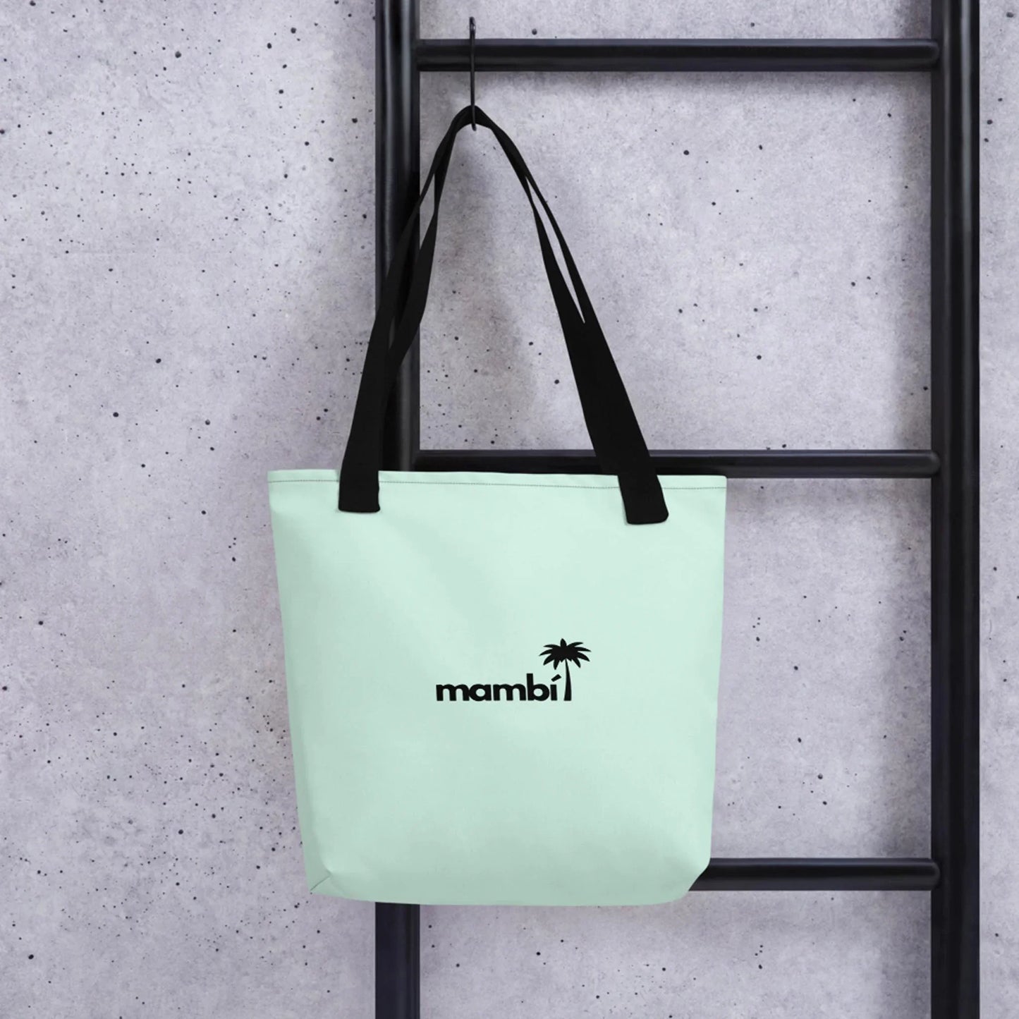 A light green Mambí tote bag with black handles hangs on a black ladder against a gray speckled wall. The bag features a small black palm tree and the word mambi printed on the front.
