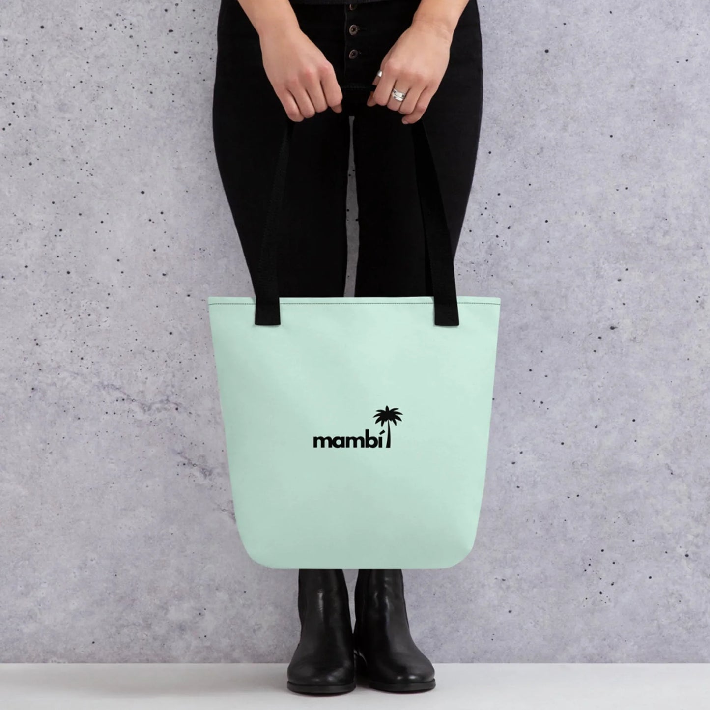A person in black jeans and boots holds a light pastel green Mambí tote bag with black straps, featuring the word mambi and a small palm tree graphic printed in black on the front.