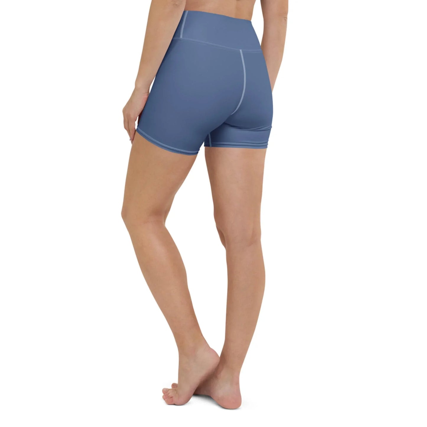 A woman stands barefoot with her back to the camera, wearing Printfuls Brickell Yoga Shorts for women—high-waisted blue shorts featuring white stitching and a supportive waistband.